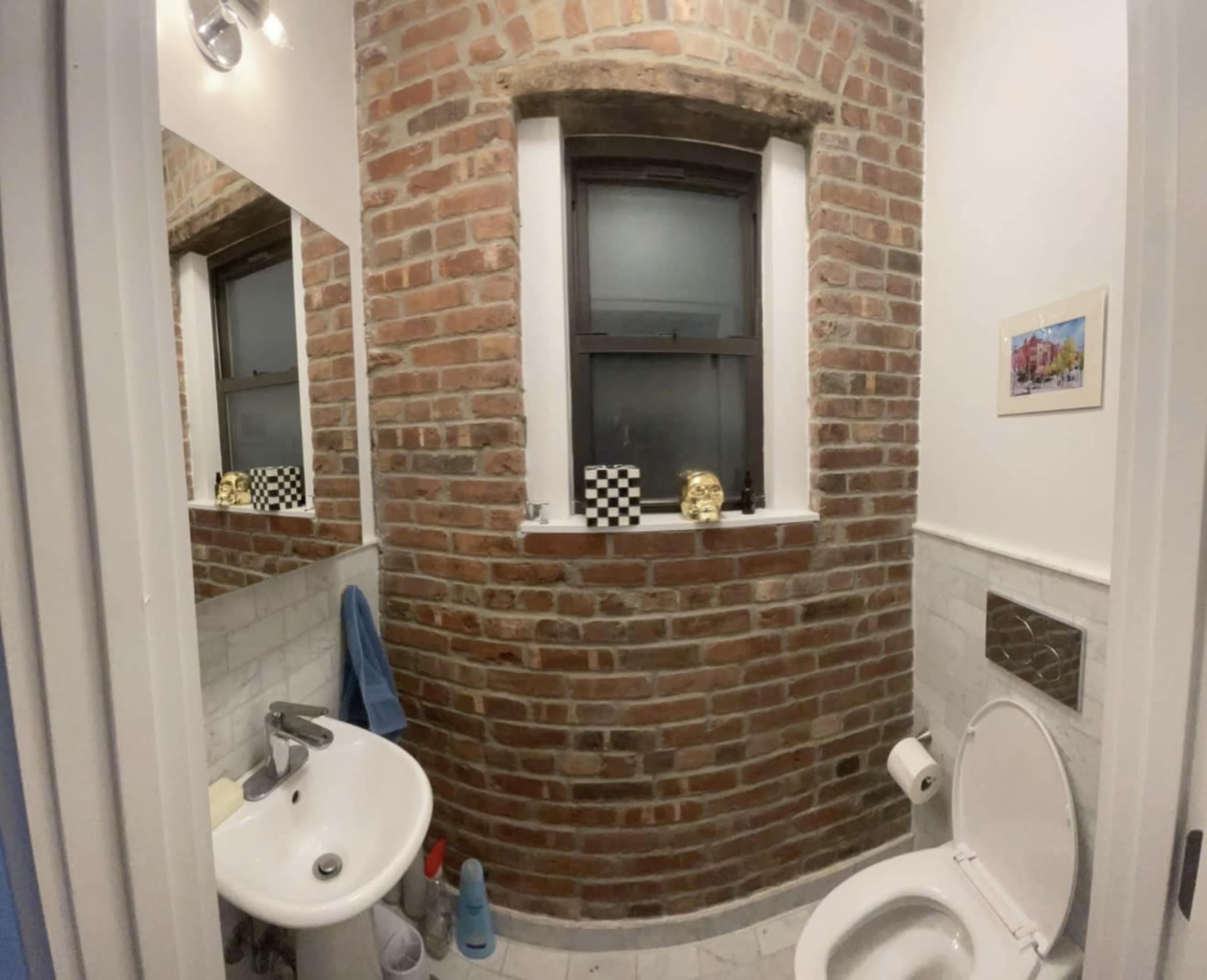 The image shows a small bathroom with exposed brick walls, a round sink, a white toilet, and a window featuring decorative items on the sill.