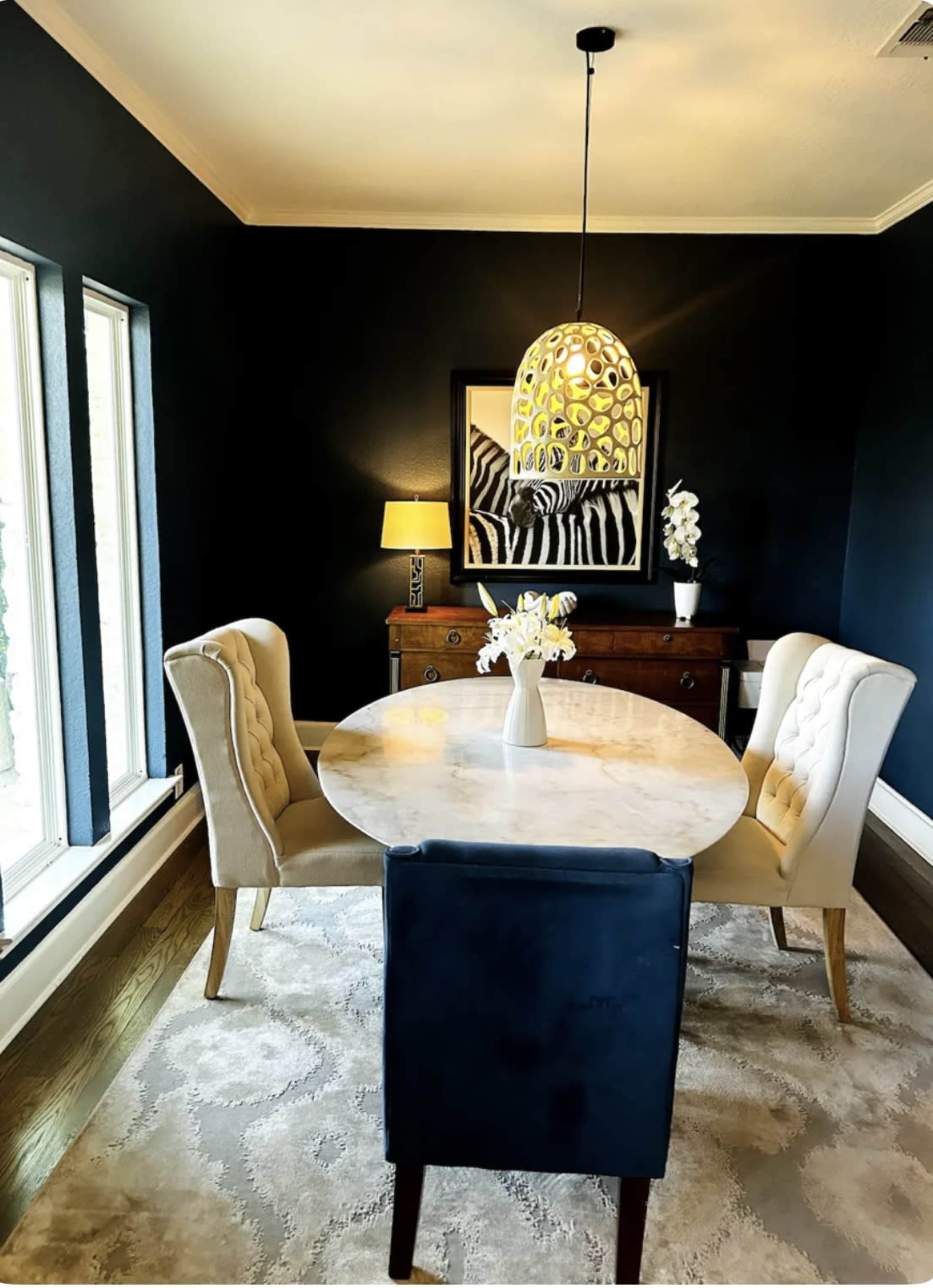 A dining area features a circular marble table surrounded by two upholstered chairs and a black chair, with a pendant light above and a side table with a lamp and artwork against a dark blue wall.