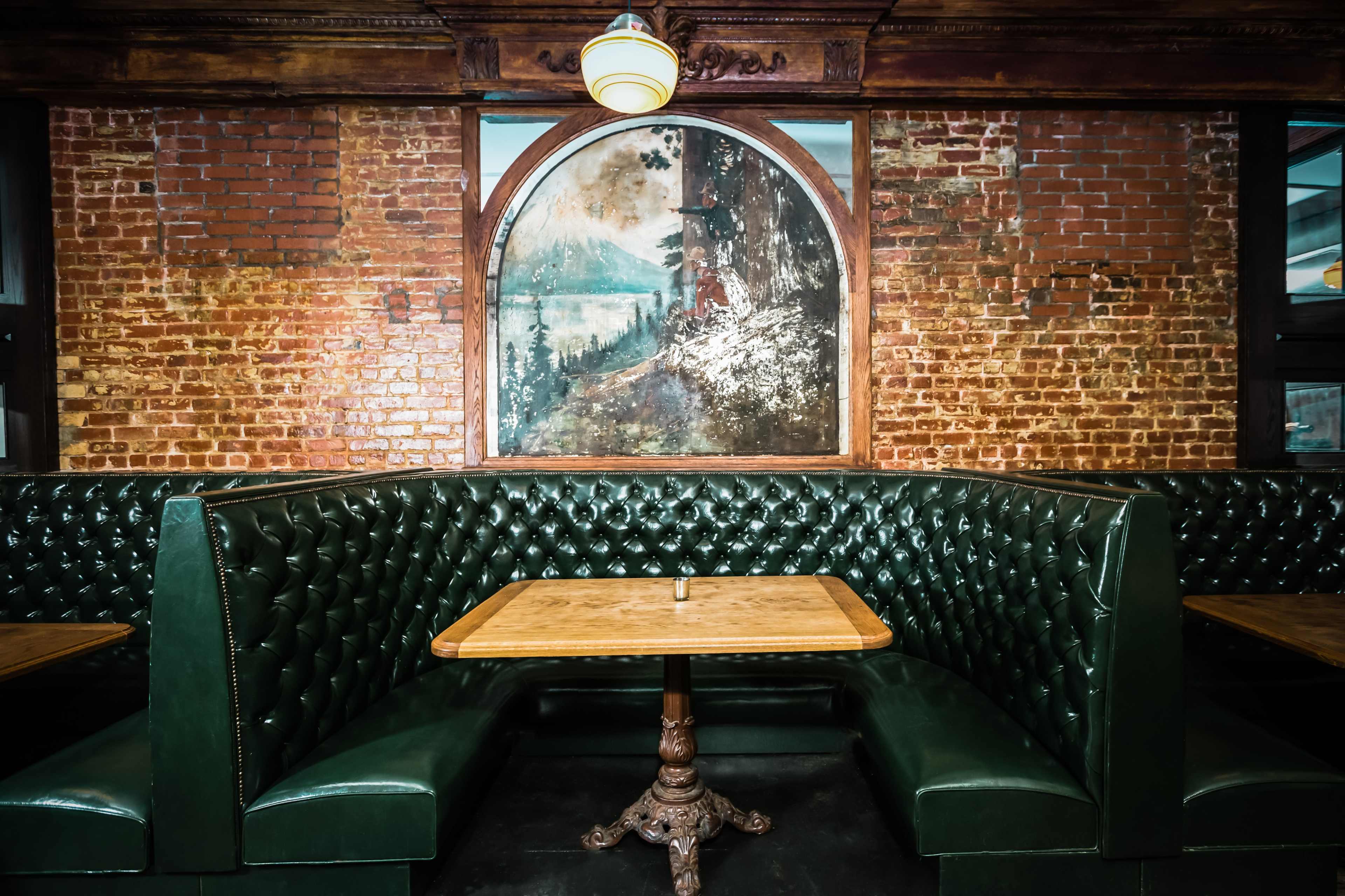 A cozy restaurant booth with dark green leather seating against a backdrop of exposed brick walls and a large framed landscape painting.