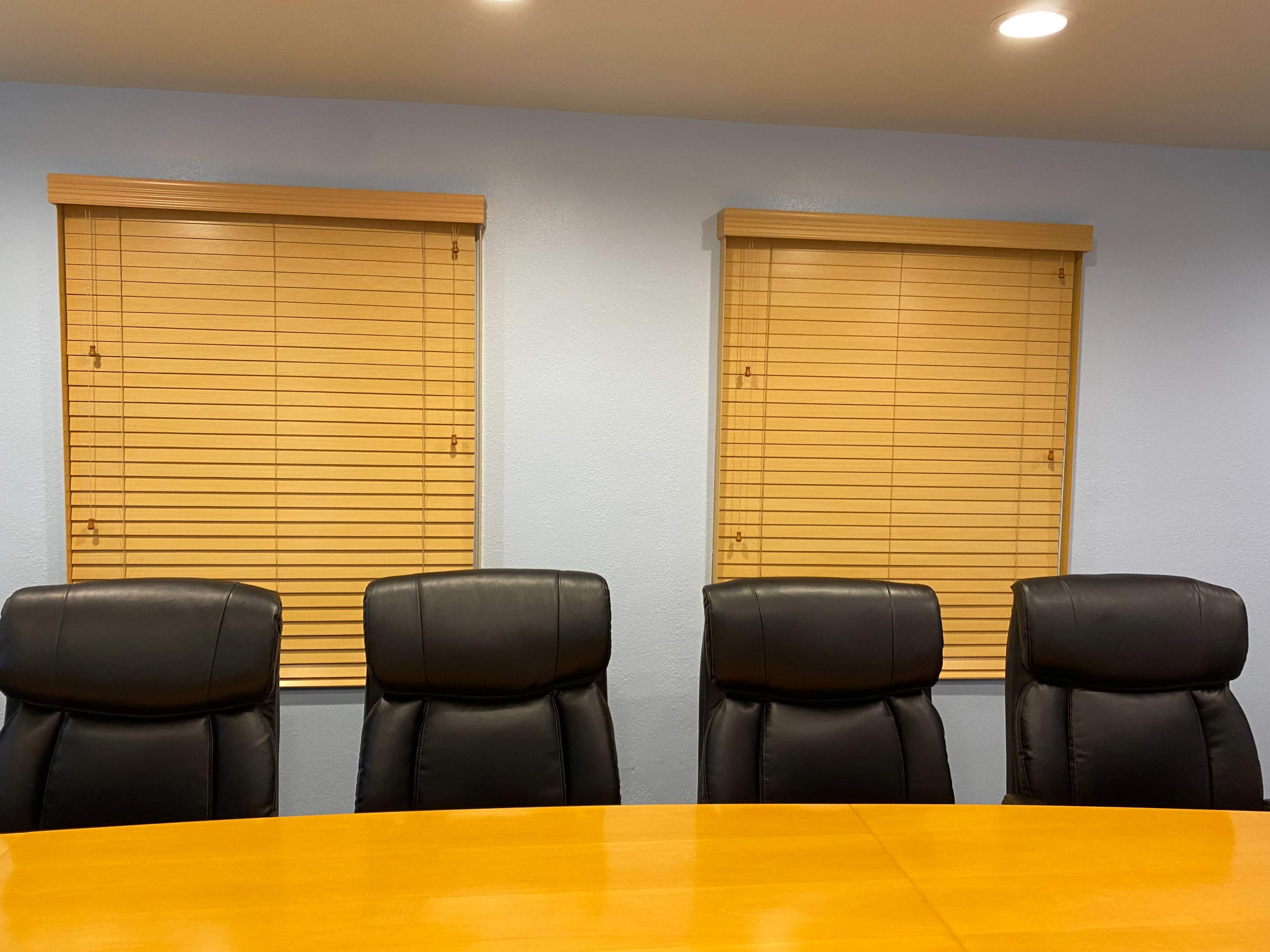 The image shows a round wooden conference table with four black leather chairs arranged around it, positioned beneath two window blinds on a light blue wall.