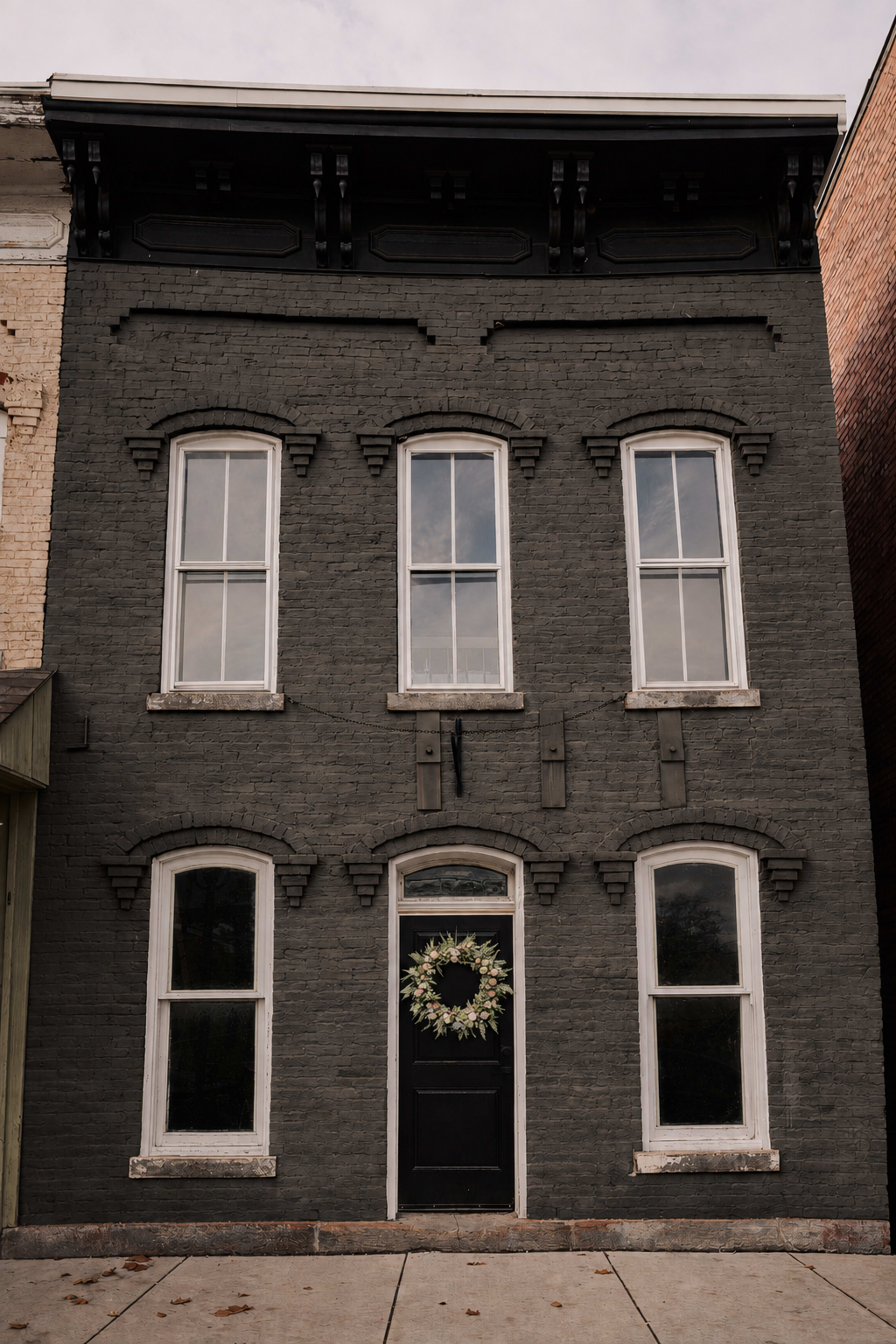 The image shows a two-story, dark gray brick building with three tall windows and a black front door adorned with a circular wreath.