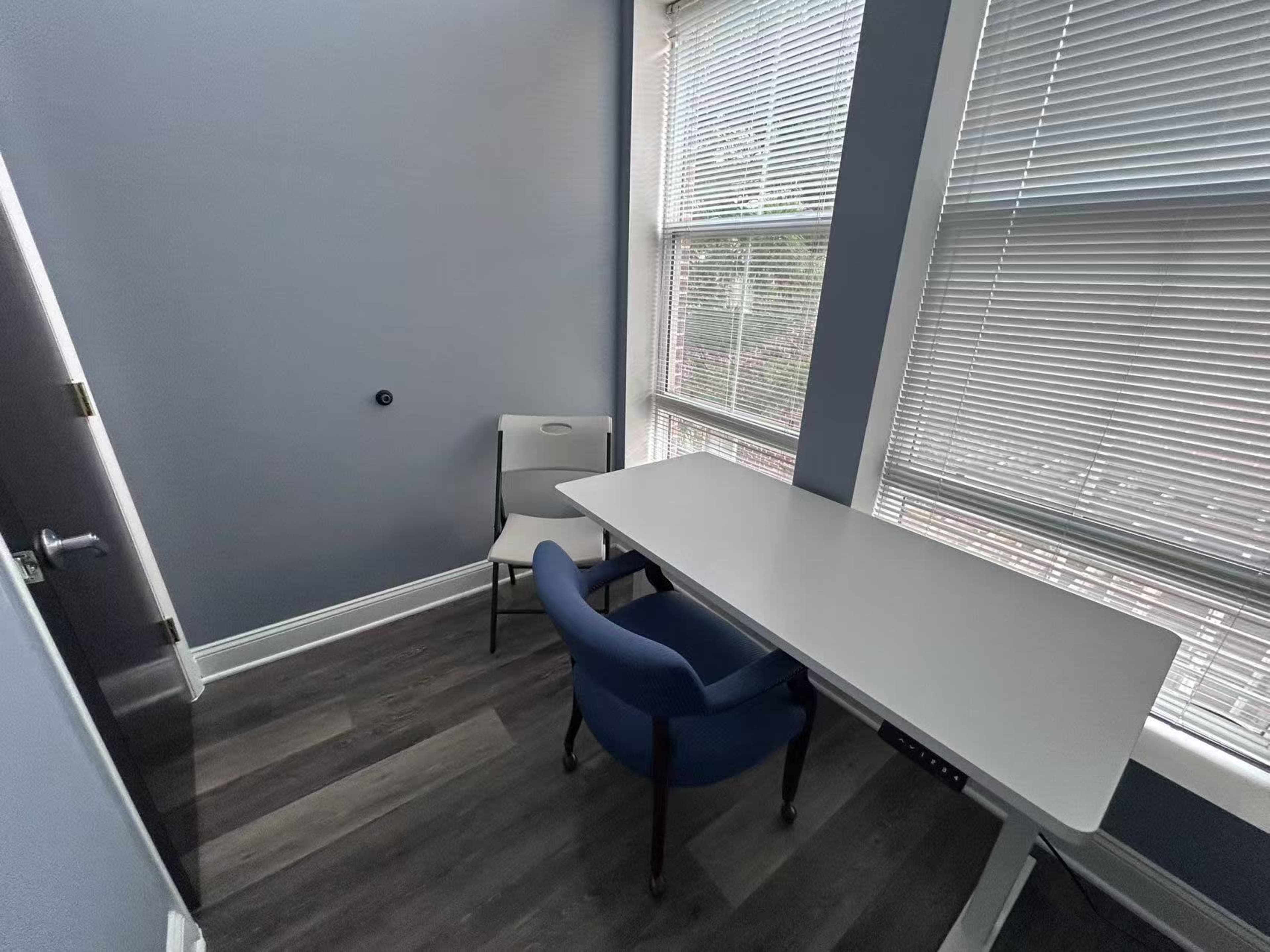 The image shows a small room with a light-colored table, a blue chair, and a folding chair near a window with blinds.