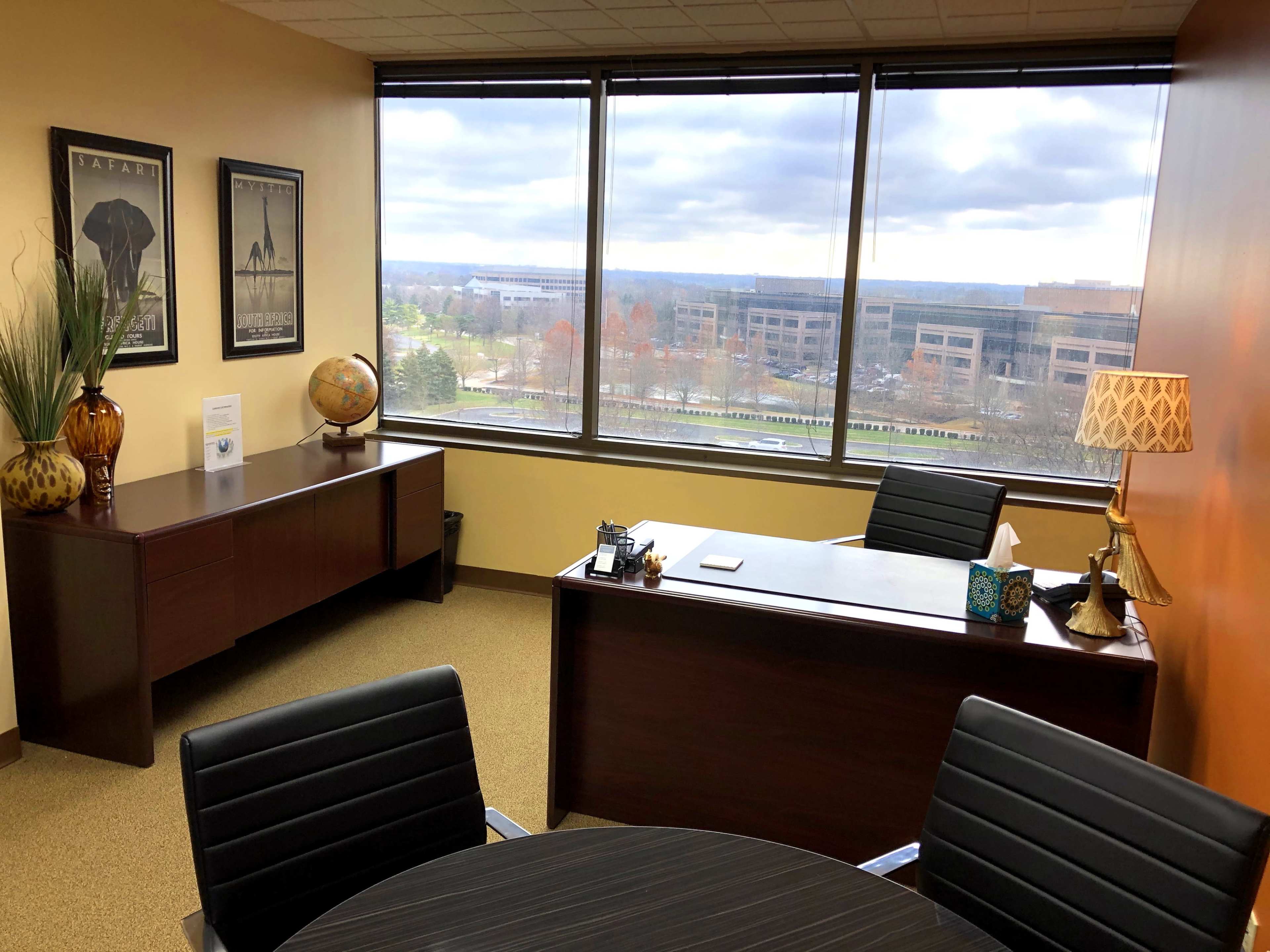 The image shows a modern office with a desk, a round table, large windows overlooking a landscape, and decorative items like a globe and a lamp.