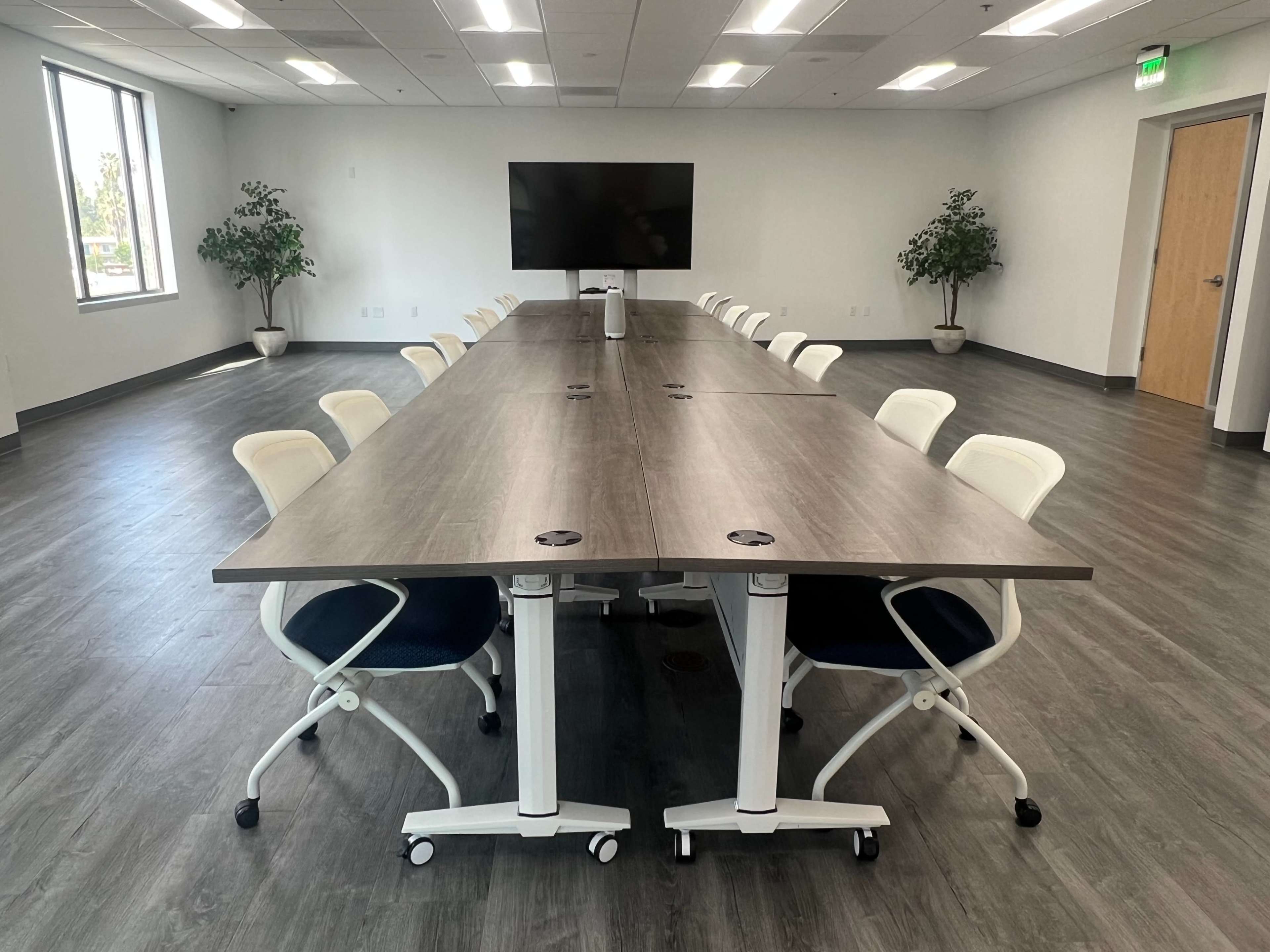 A spacious conference room with a long, rectangular table surrounded by white ergonomic chairs, and a large screen mounted on the wall.
