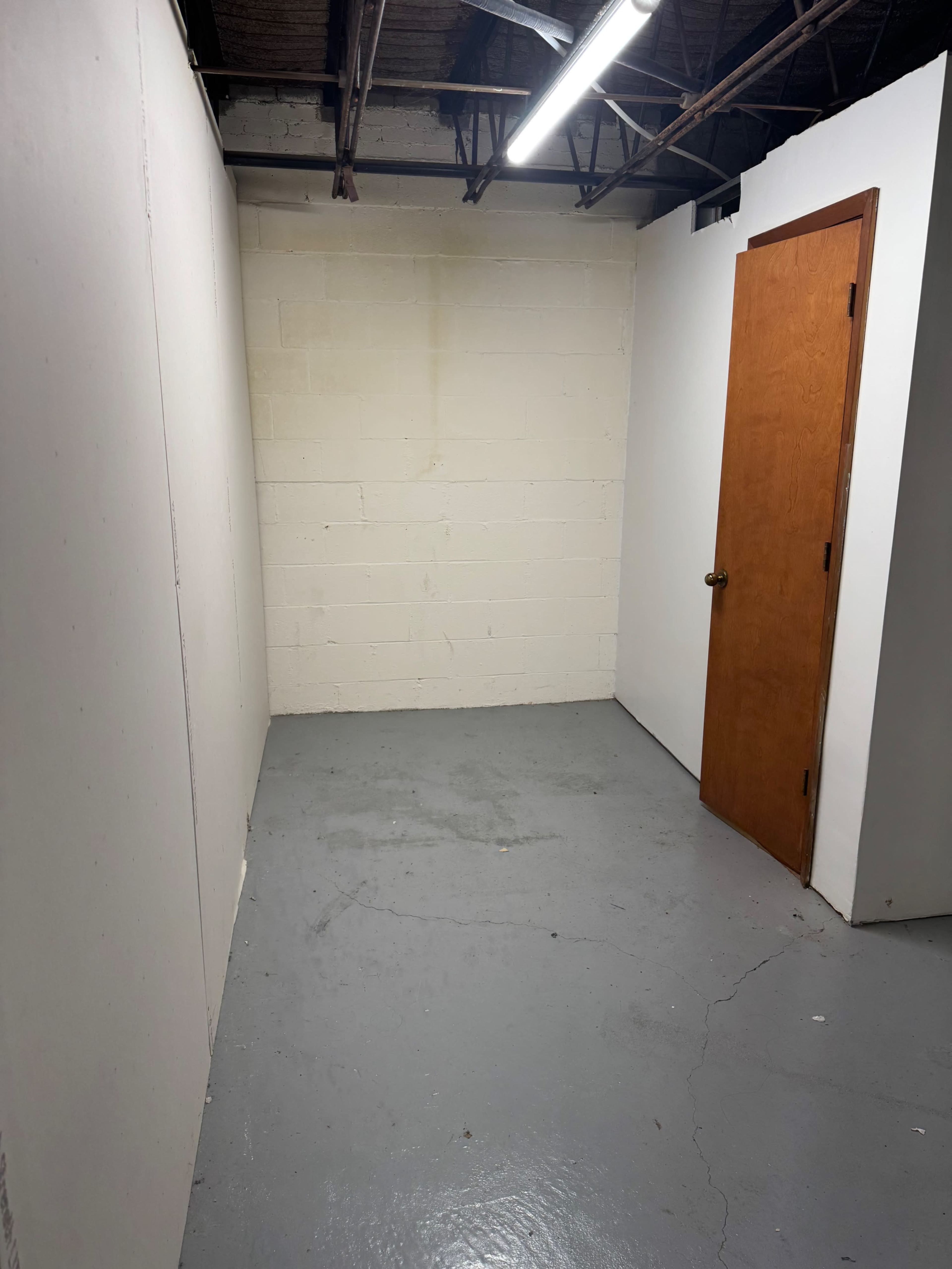 The image shows a small, empty room with concrete flooring, bare walls, and a wooden door on one side.