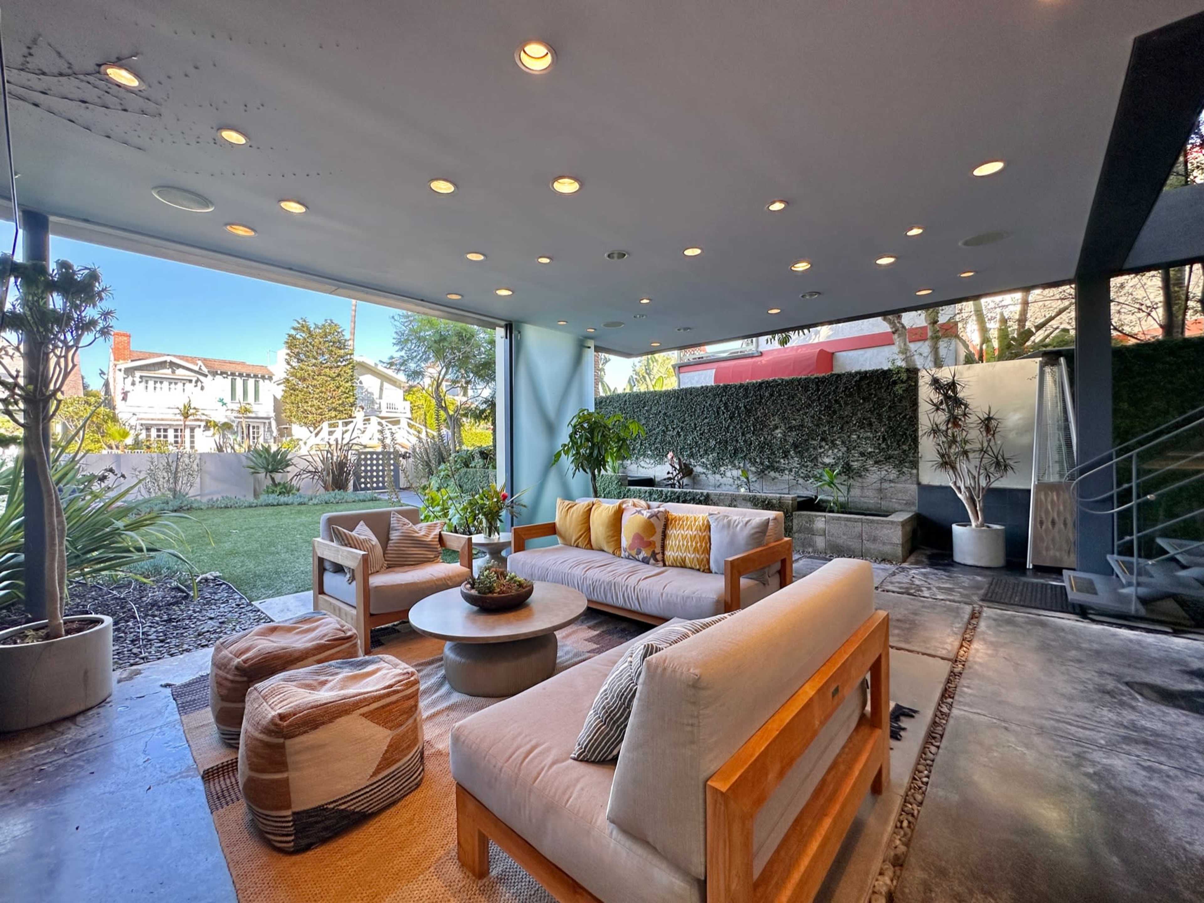 A modern outdoor living space with a seating area featuring wooden furniture, cushions, and a small garden visible through large glass doors.