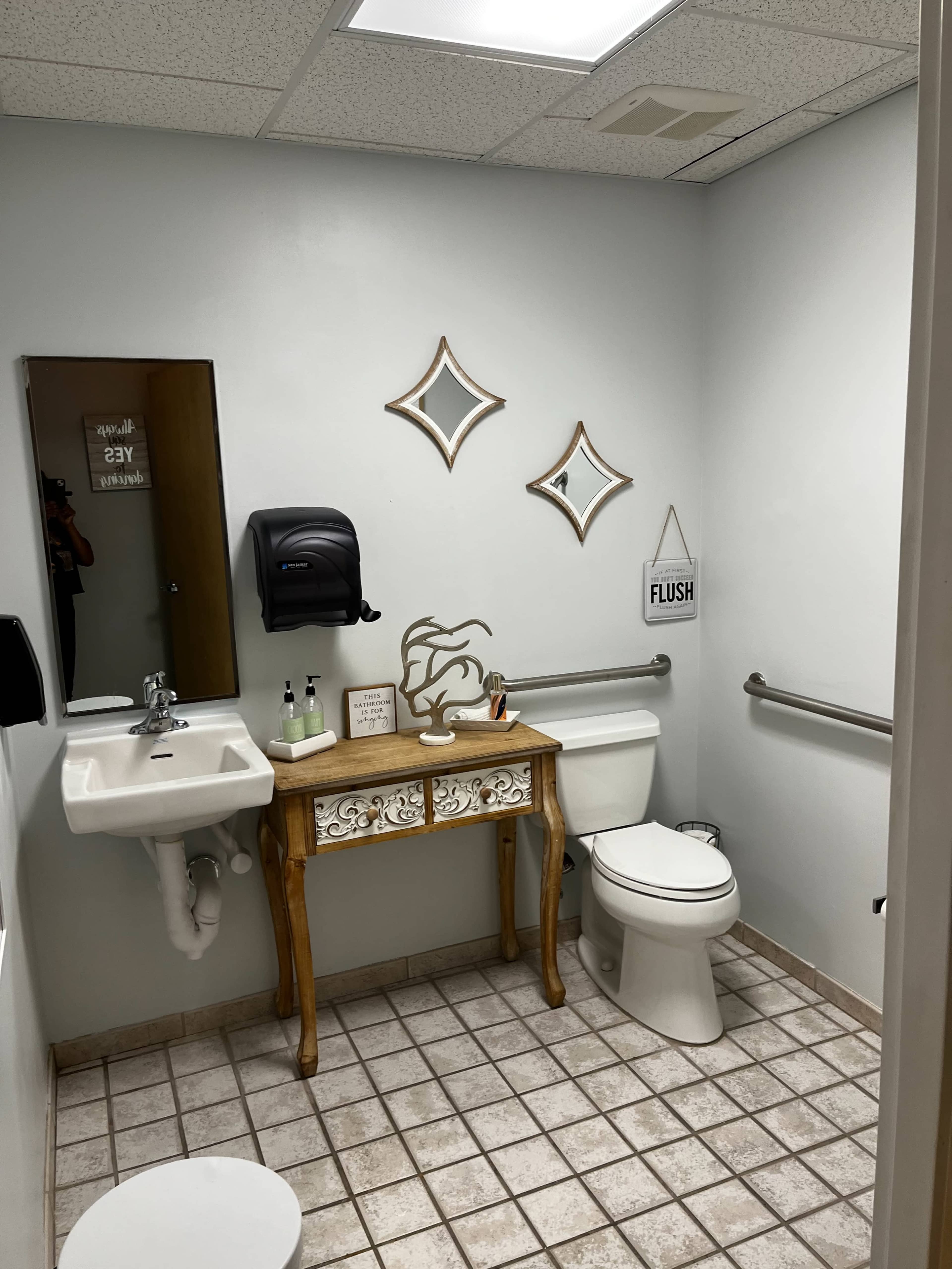A restroom featuring a toilet, a sink with a mirror above it, decorative wall mirrors, and a small wooden table with various items on it.