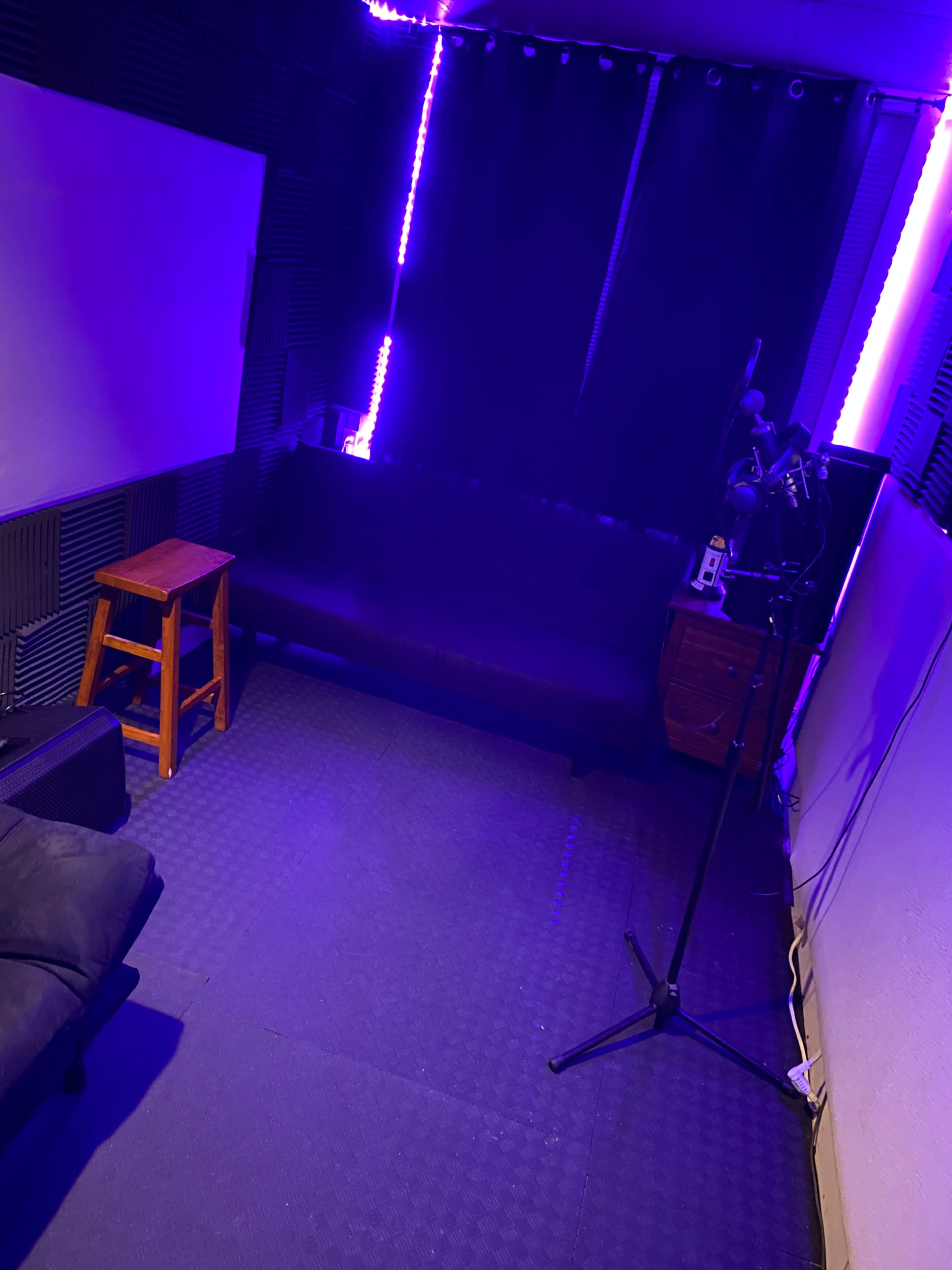 A dimly lit sound studio with purple LED lights, a black couch, a stool, and a microphone stand.