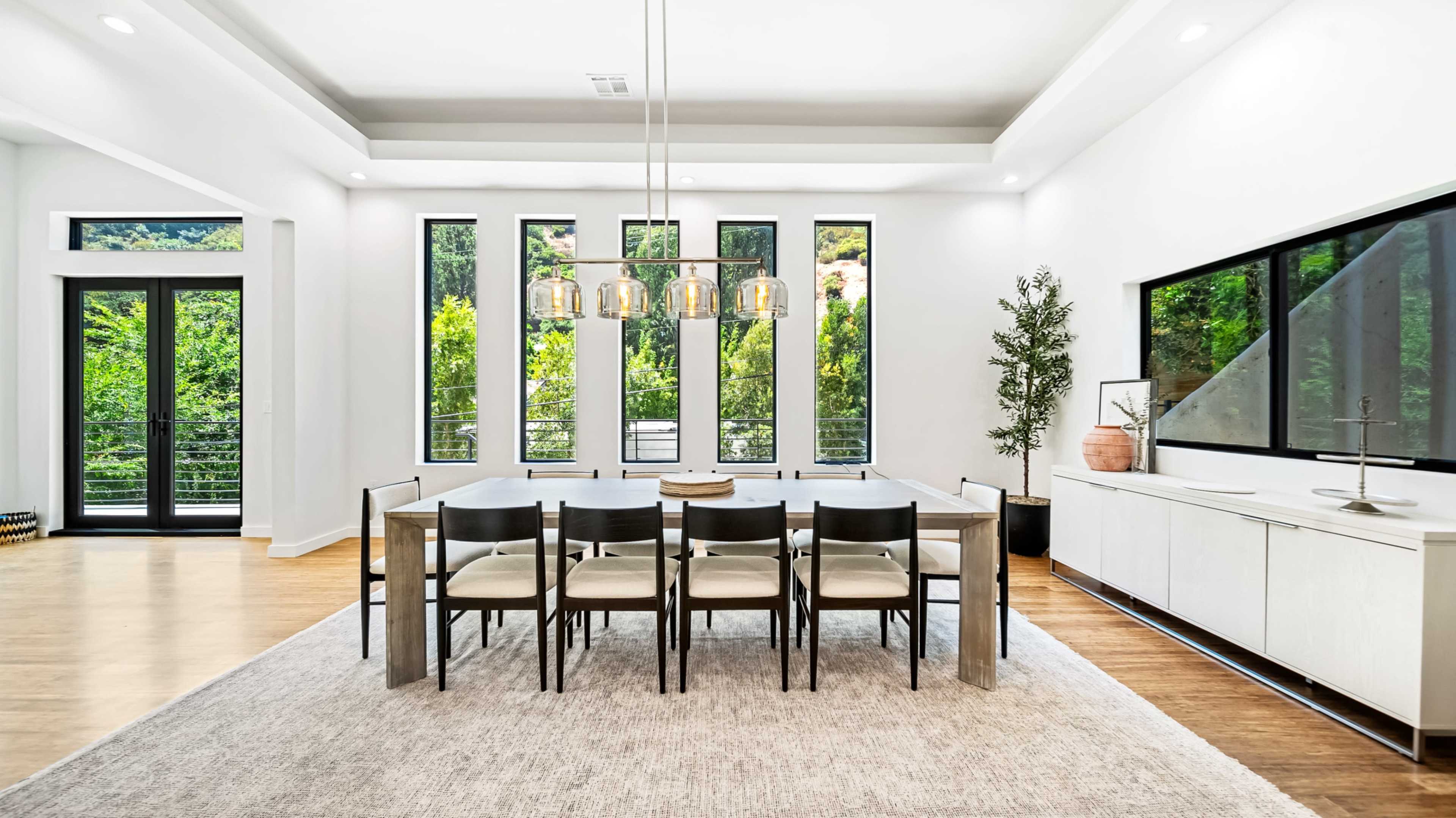 A modern dining room features a long table with eight chairs, surrounded by large windows that offer a view of greenery outside.