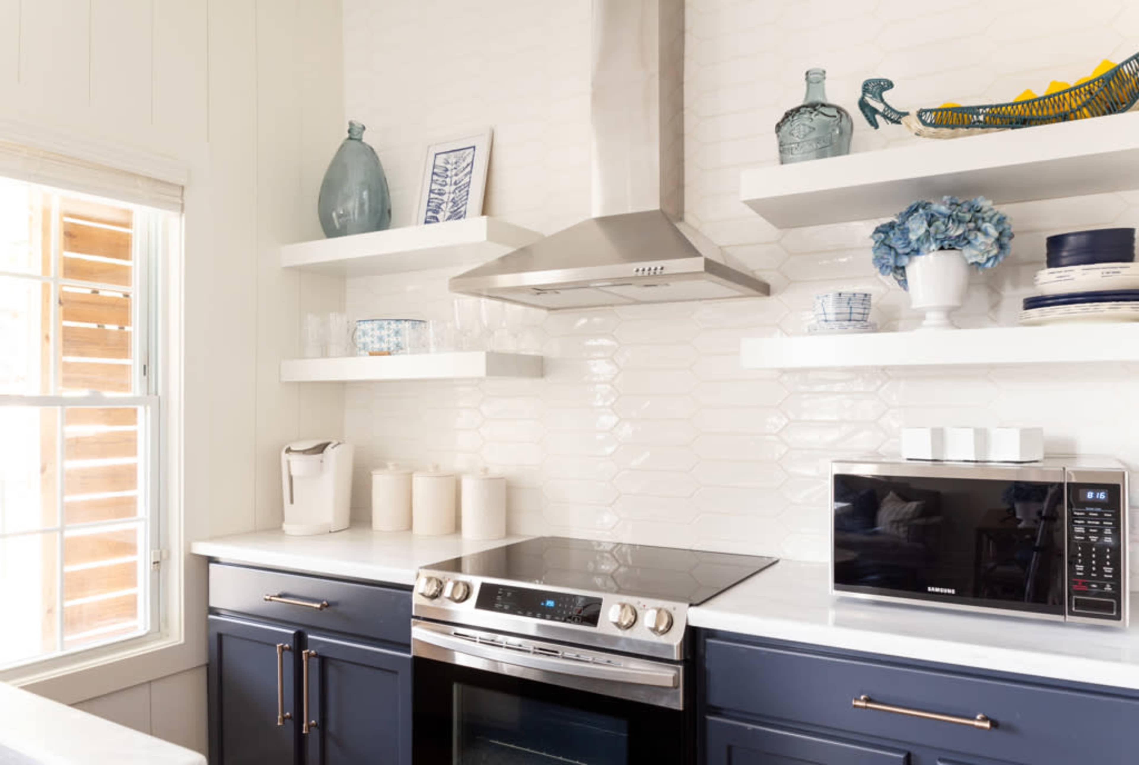 A modern kitchen features a stainless steel stove and hood, white tile backsplash, and open shelving with decorative items.