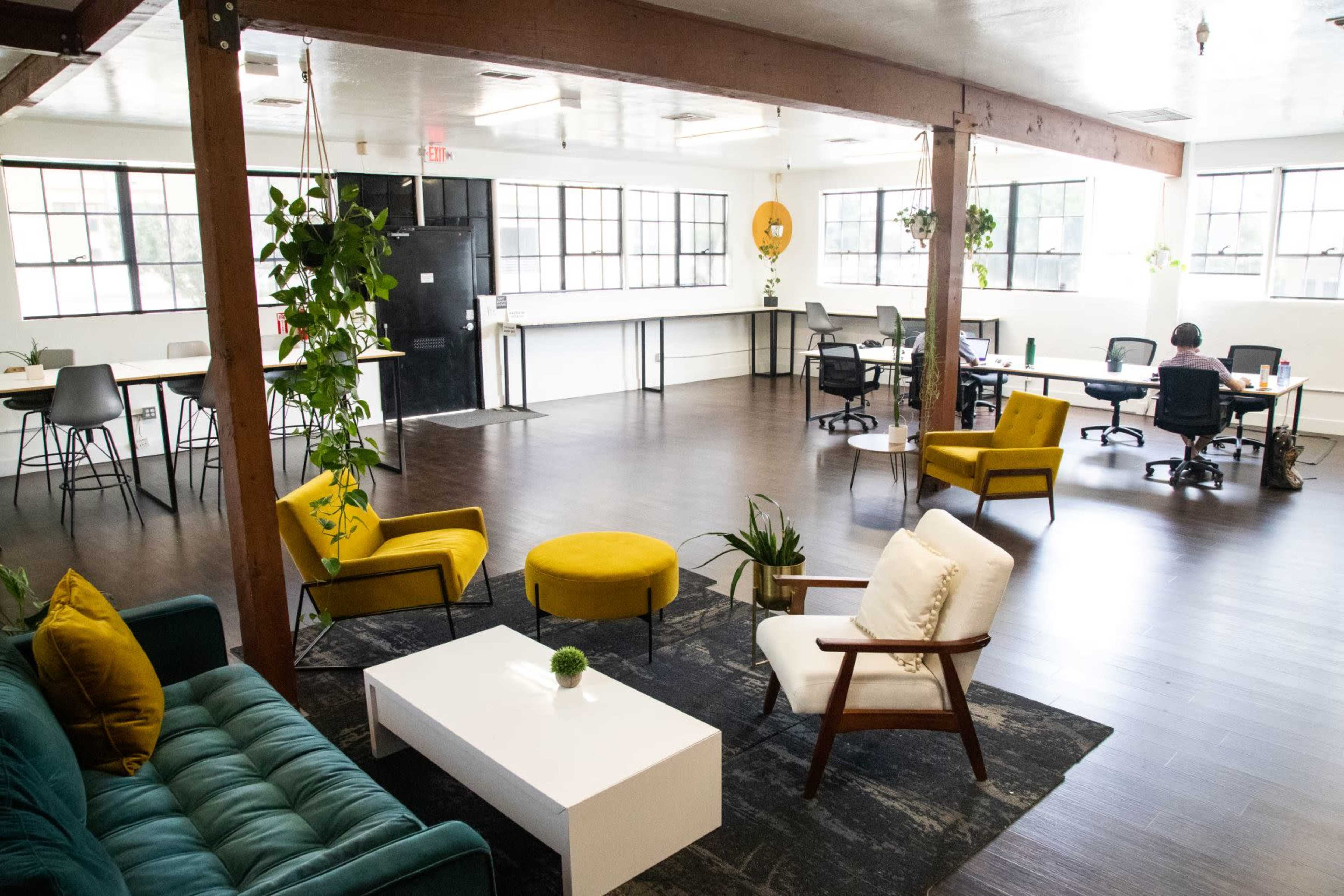 A modern office space featuring seating areas with chairs and a sofa, large windows, and several workstations.