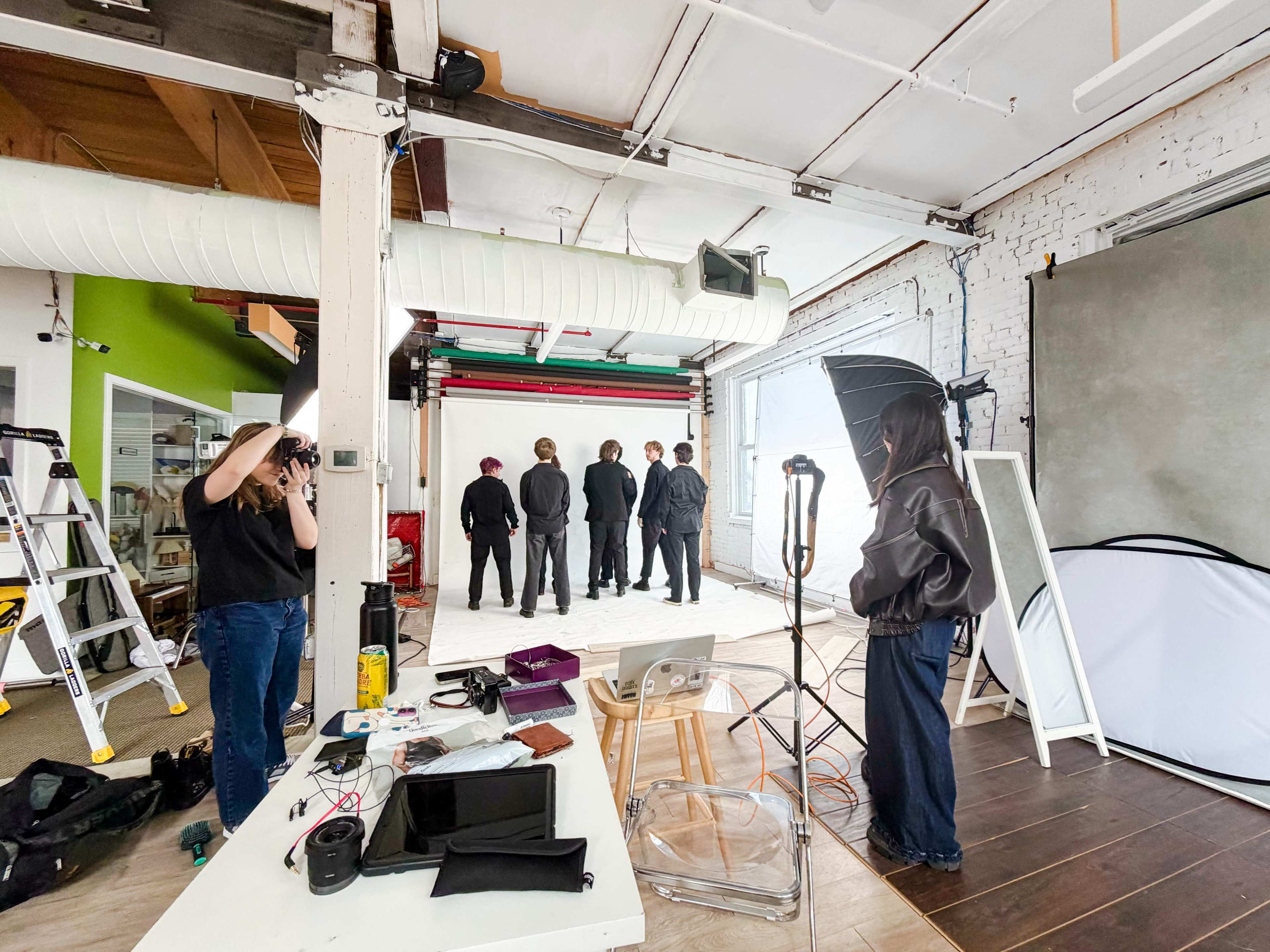 A photography studio features a group of people dressed in black, standing in the background, while two individuals are preparing equipment in the foreground.