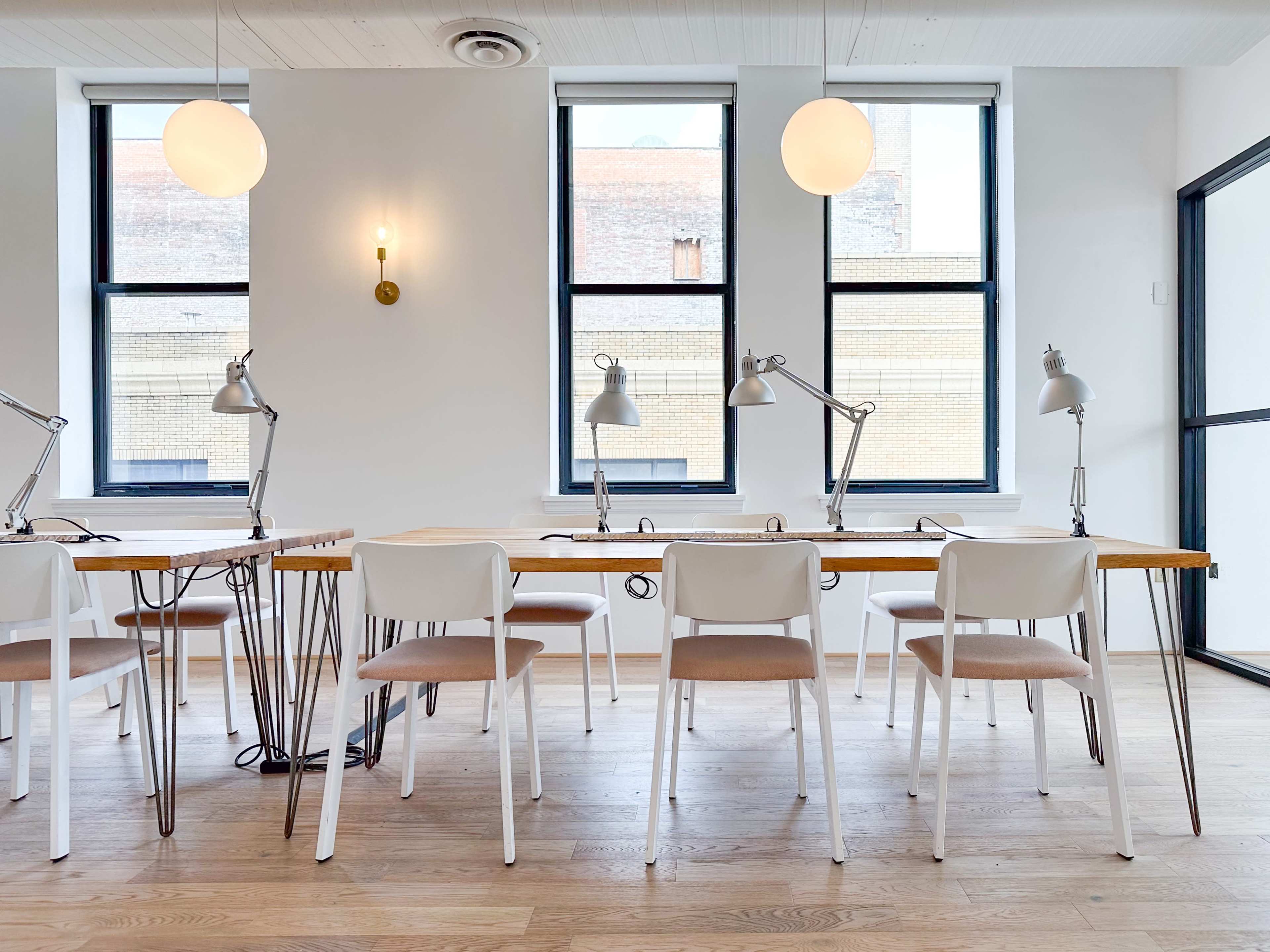 A bright, modern workspace with several white desks and task lamps arranged along a wall of large windows.