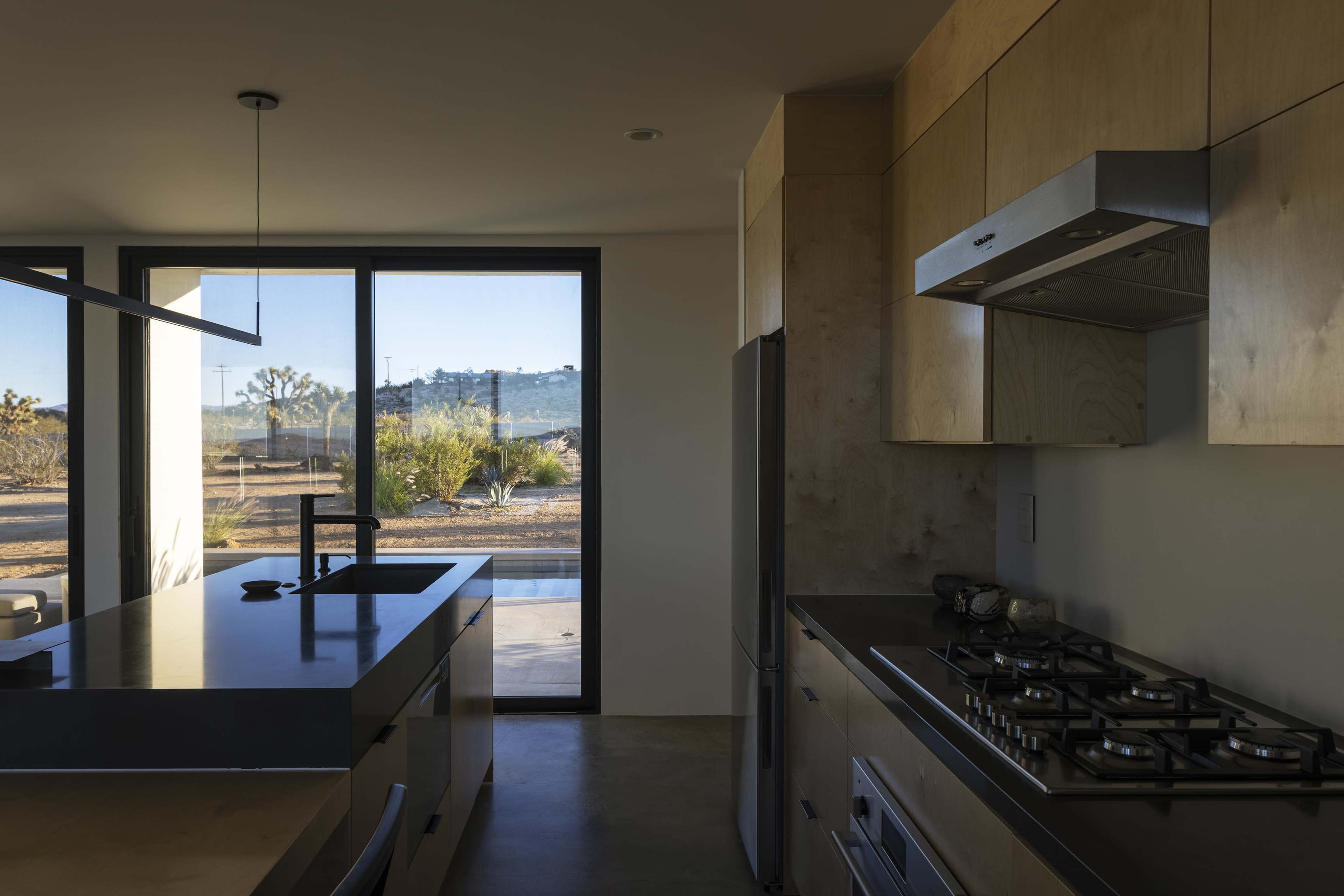 A modern kitchen with a stainless steel stove and wooden cabinetry, featuring a large window that overlooks a desert landscape.