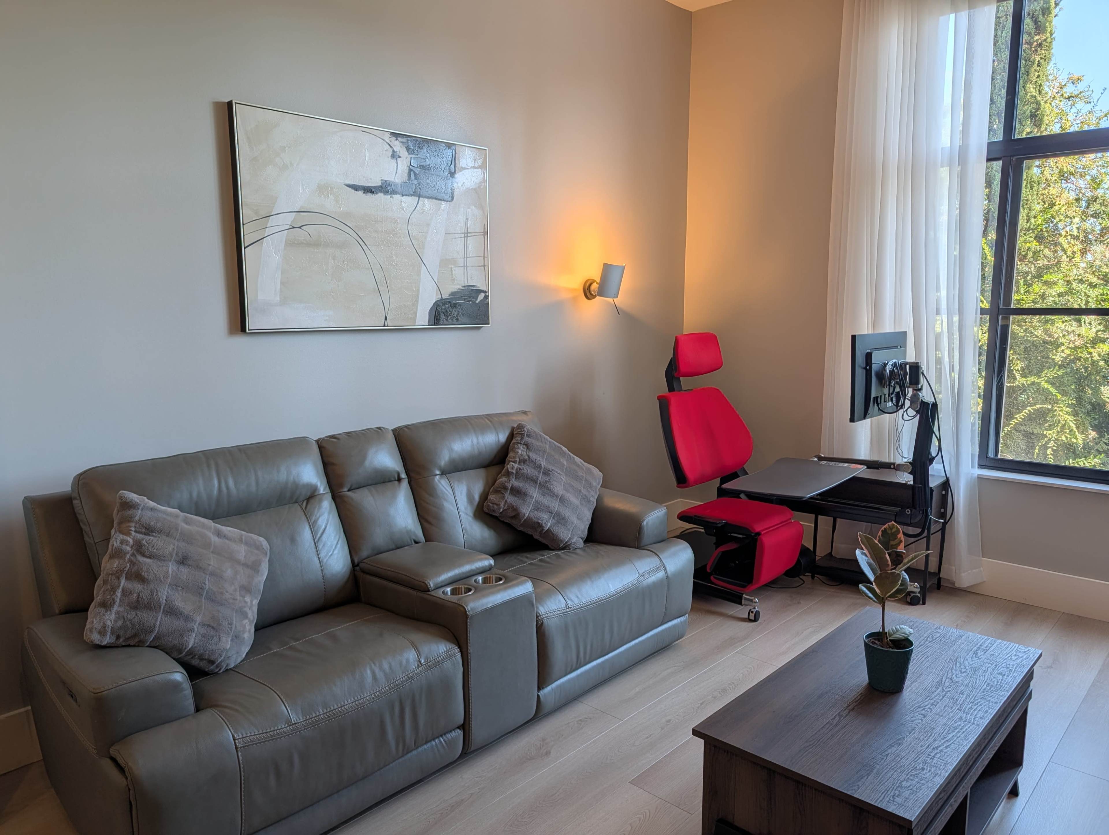 The image shows a modern living room featuring a gray leather sofa with decorative pillows, a wooden coffee table, a red ergonomic chair, and a computer setup beside a large window.