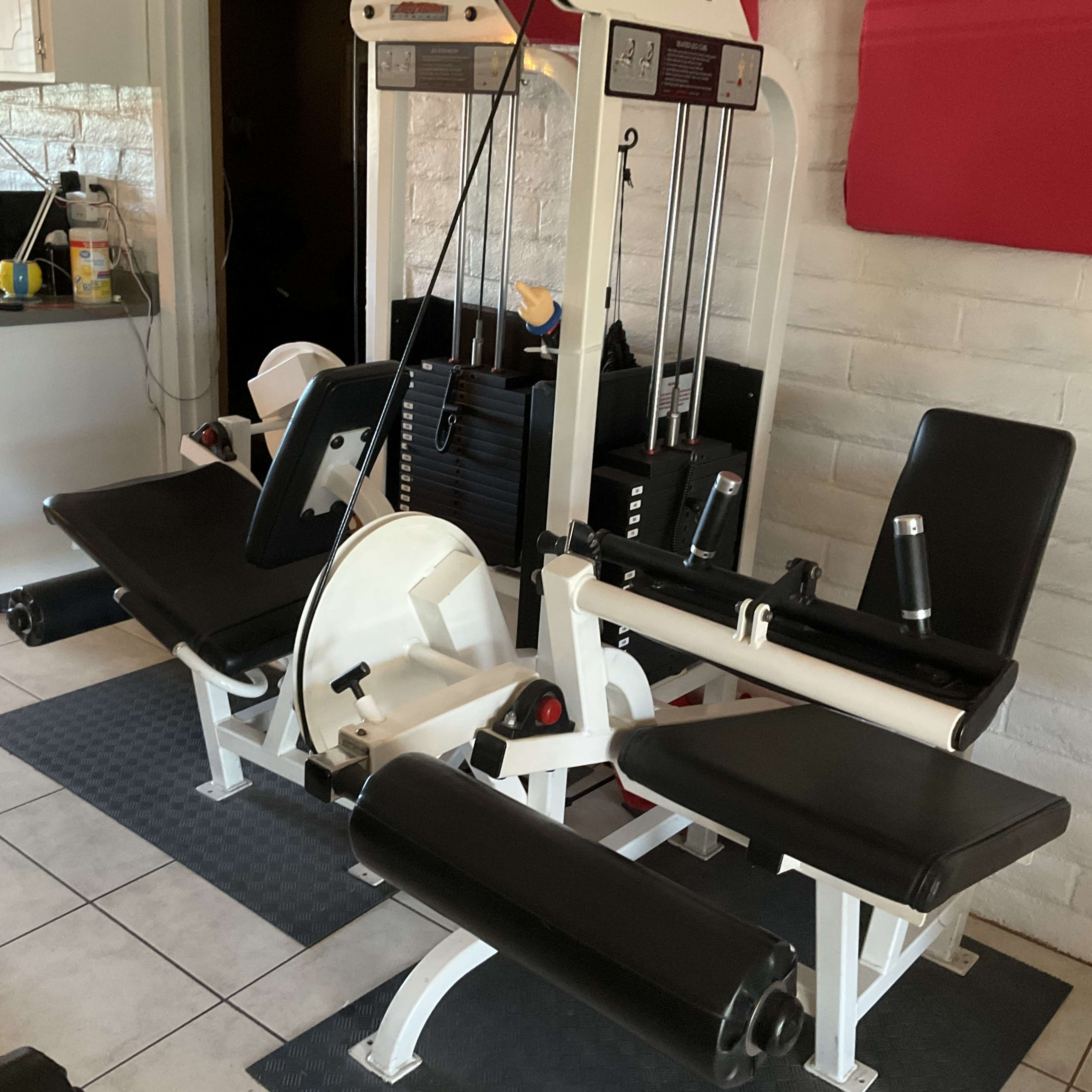 The image shows a multi-station gym machine with various exercise apparatus, including leg and arm components, set in a room with tiled flooring.