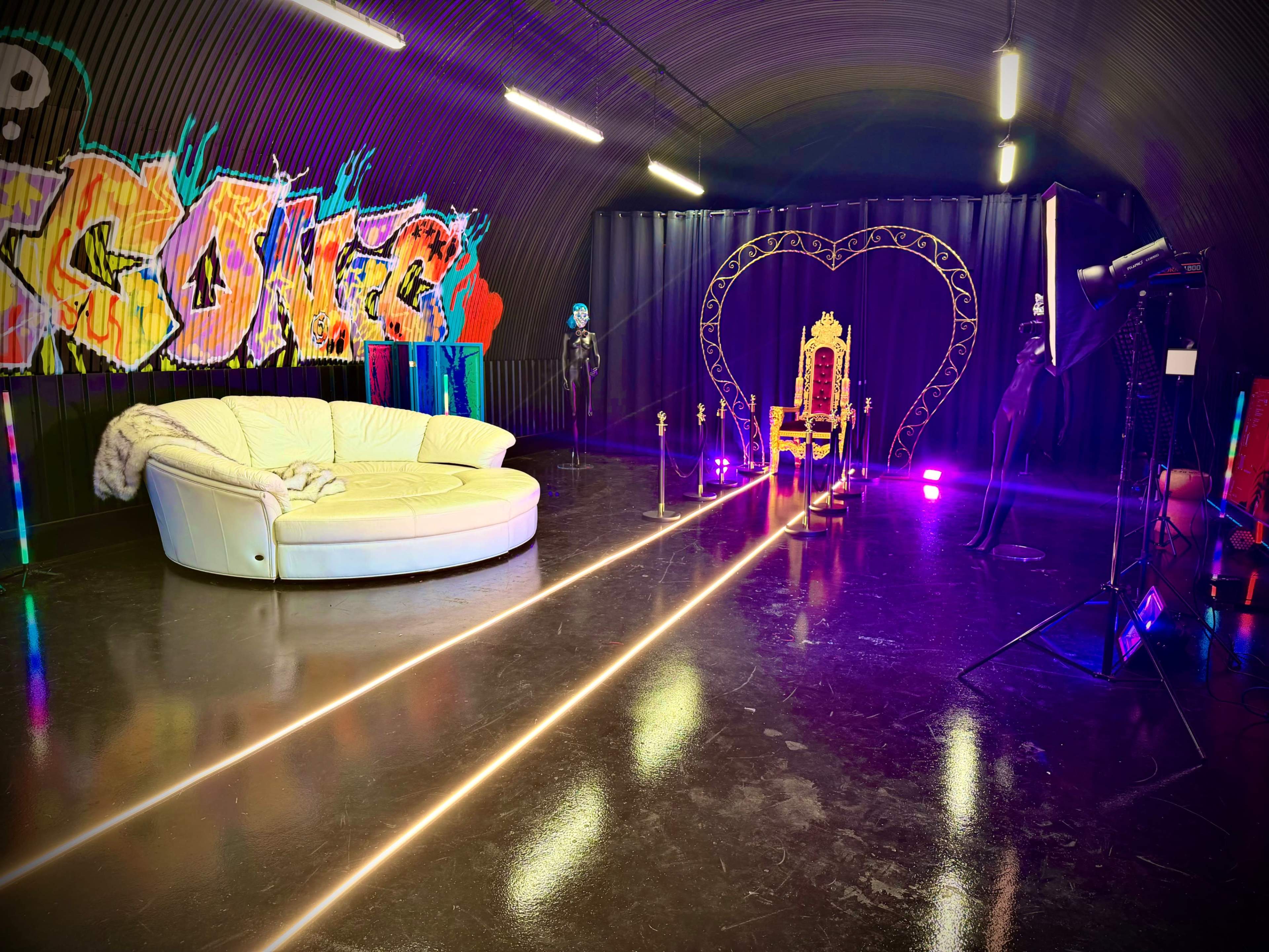 The image depicts a modern studio space featuring a white circular couch, a decorative throne, and vibrant graffiti artwork, all illuminated by colorful lights.