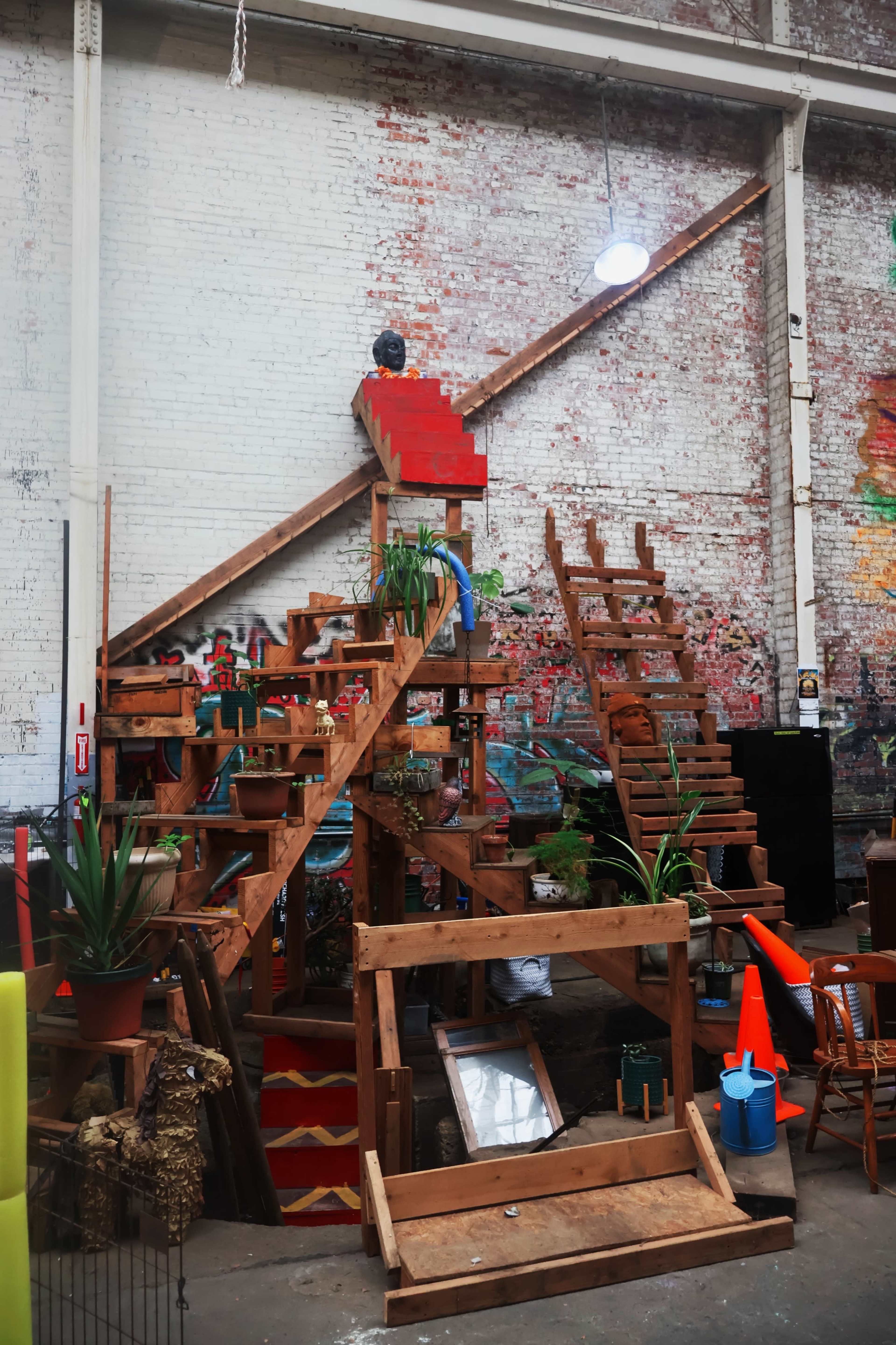 The image shows a large wooden structure with multiple staircases, planted greenery, and various items placed around in an industrial space with graffiti on the walls.