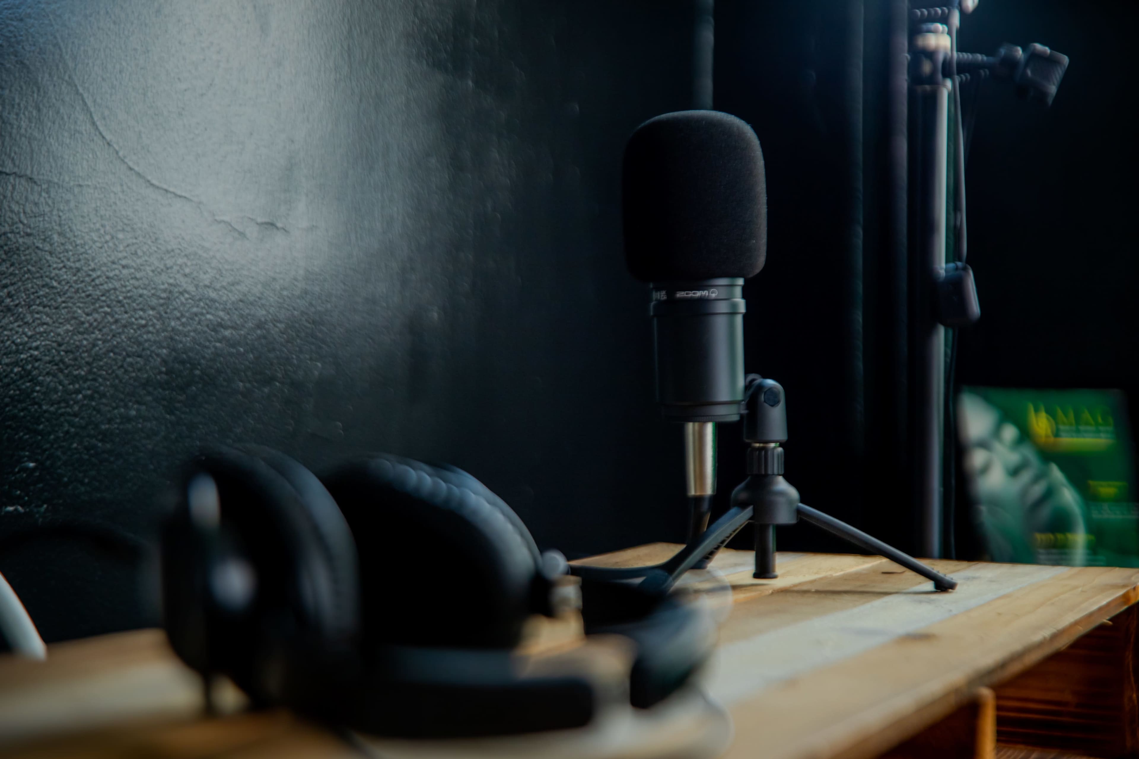 A microphone sits on a stand next to a pair of headphones on a wooden surface in front of a dark wall.