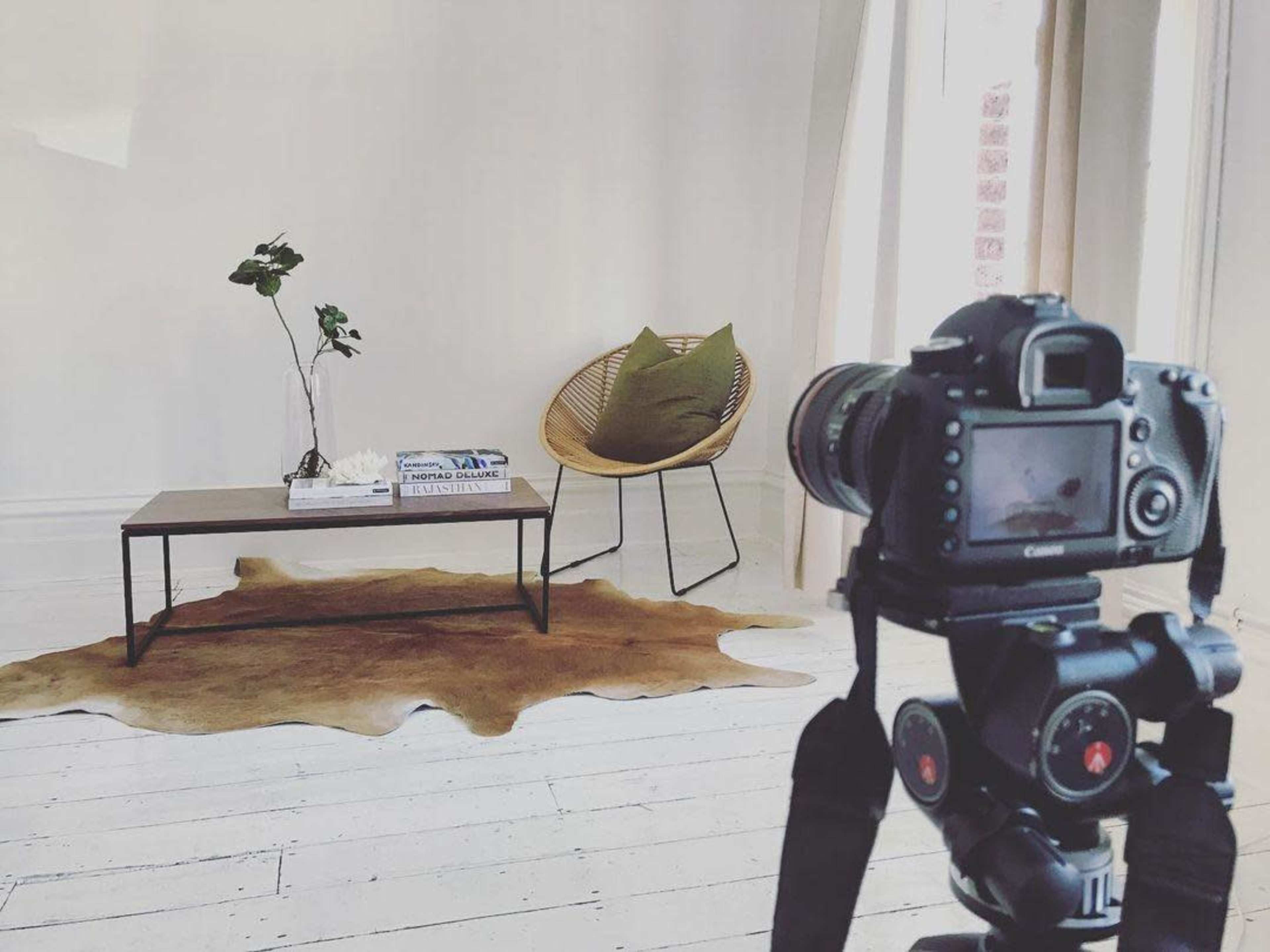 A camera is focused on a minimalist living room scene featuring a wooden coffee table, a plant, a decorative object, and a chair on a cowhide rug.