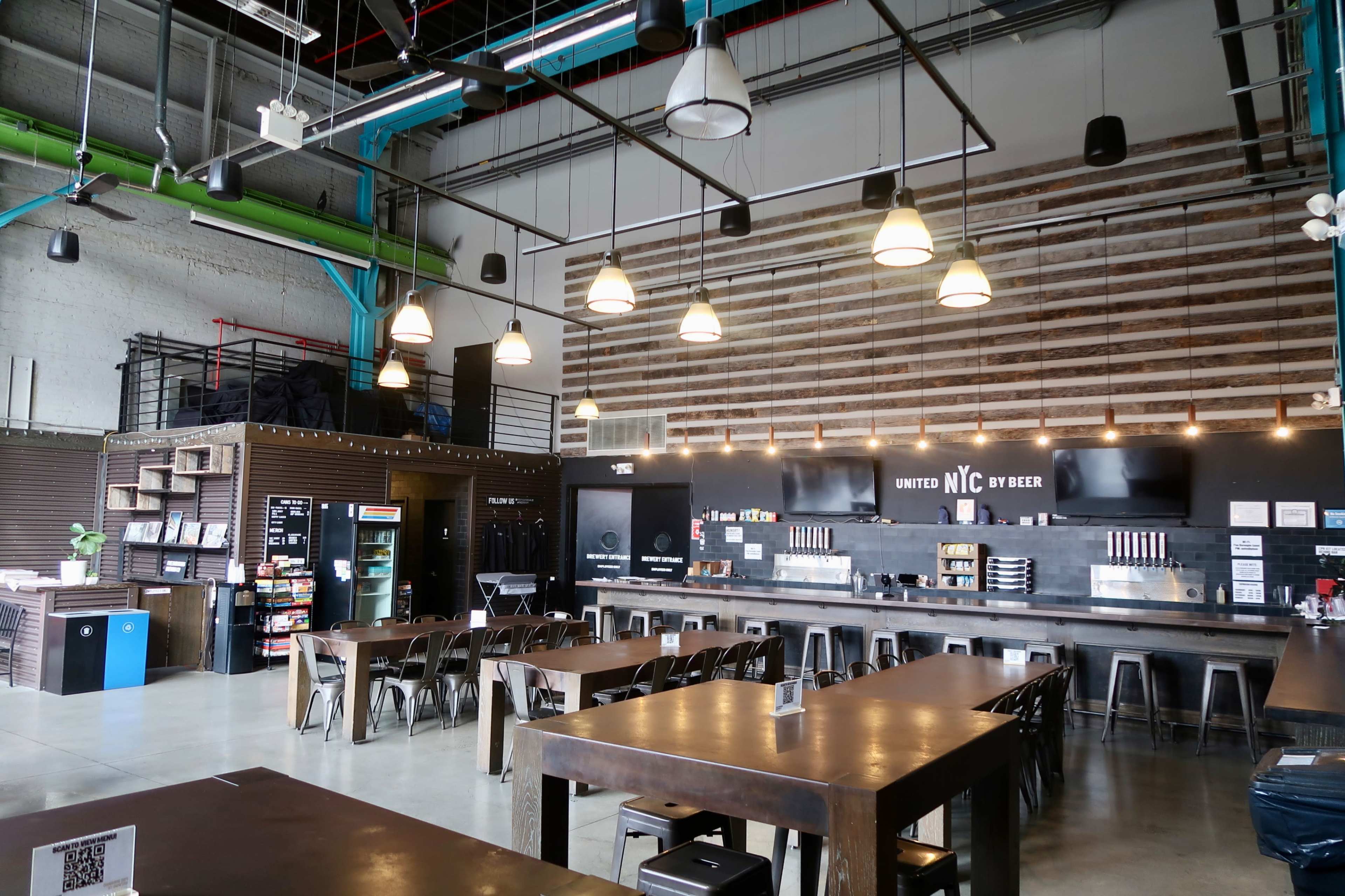 The image shows a spacious, modern café with long wooden tables, industrial lighting, and a coffee bar featuring various brewing equipment and a menu board.