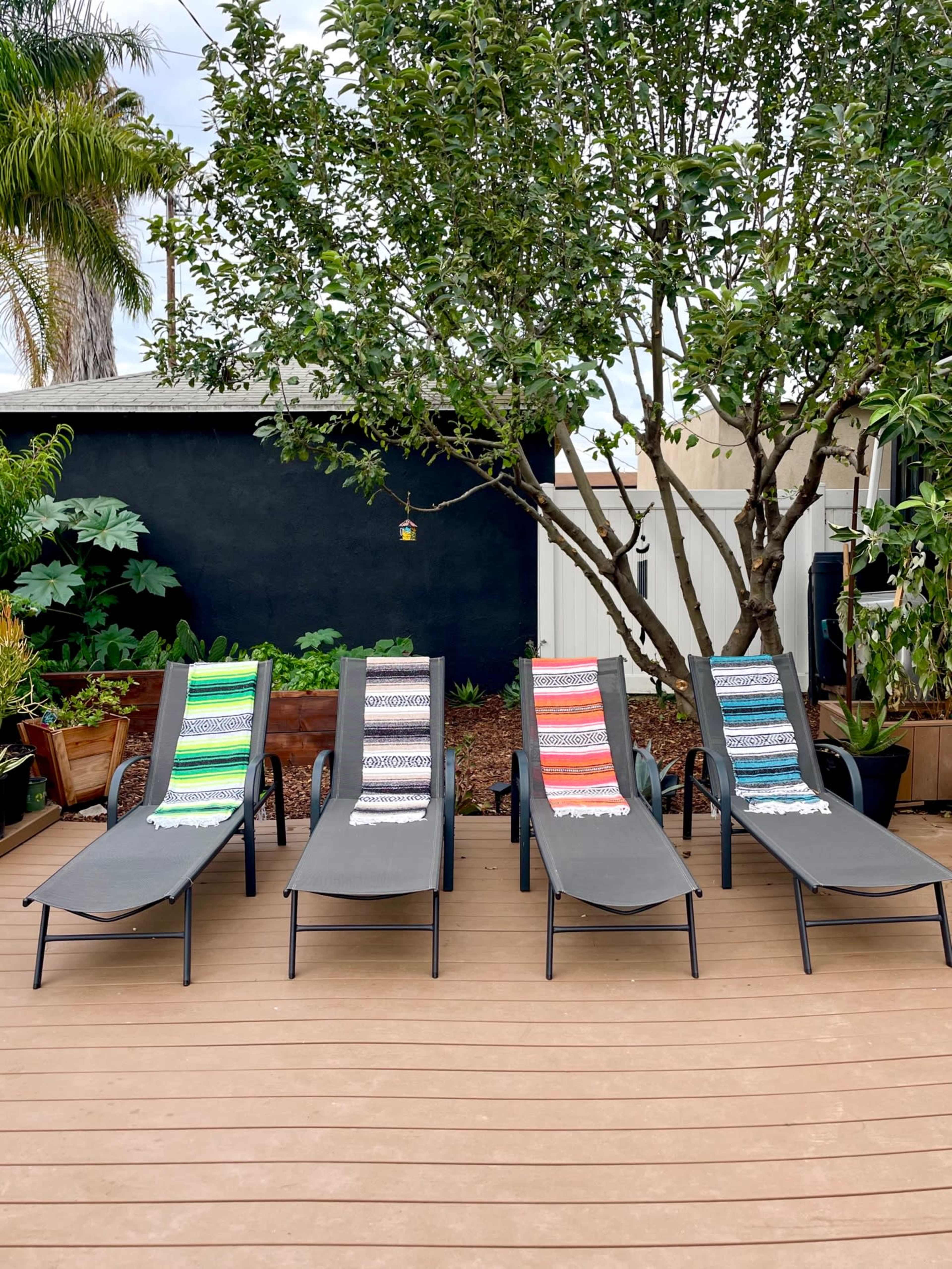 A row of four lounge chairs with colorful woven blankets sits on a wooden deck surrounded by greenery.