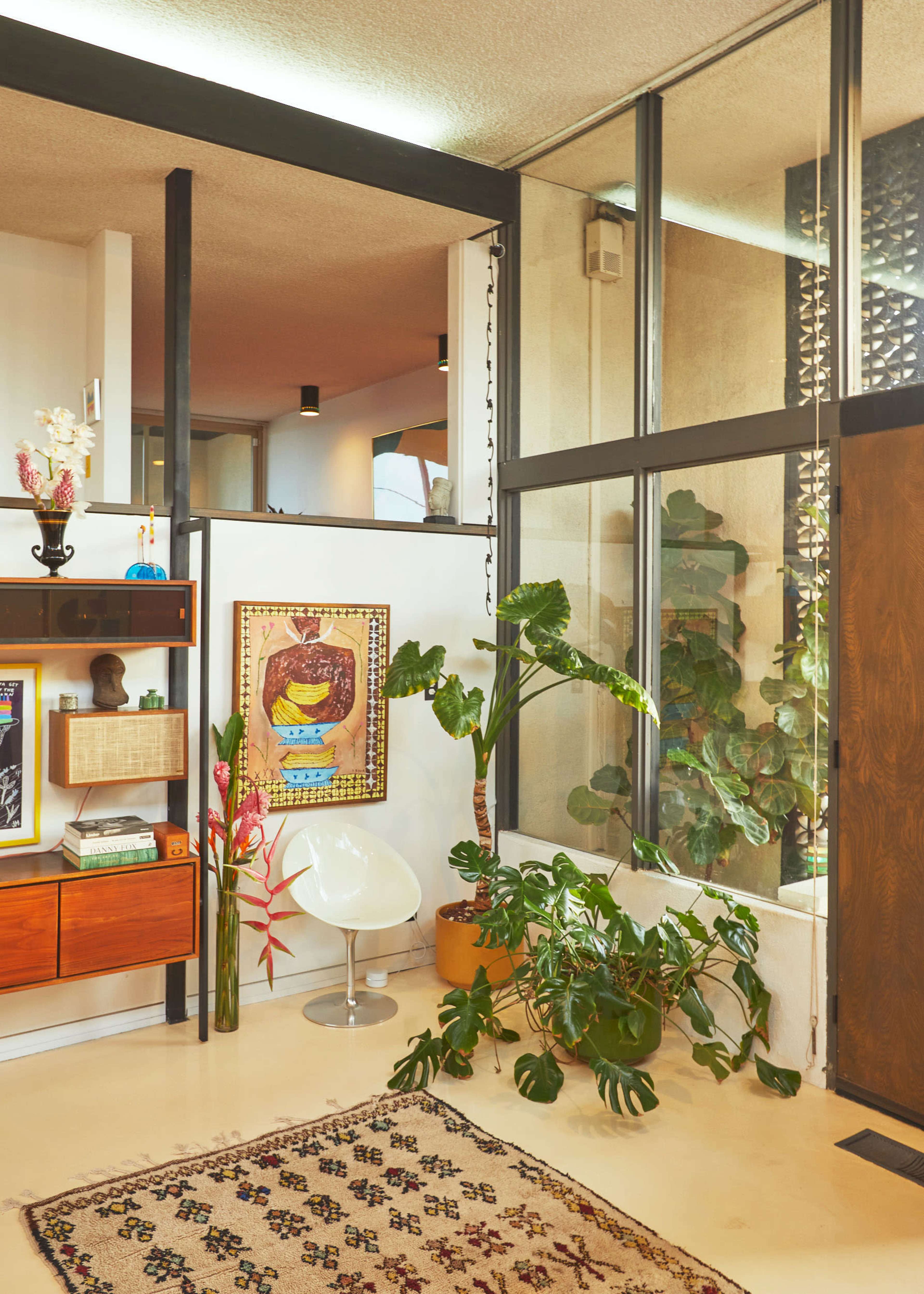 A modern interior space with a wooden shelf, a white chair, a large potted plant, and a patterned rug near a glass wall.