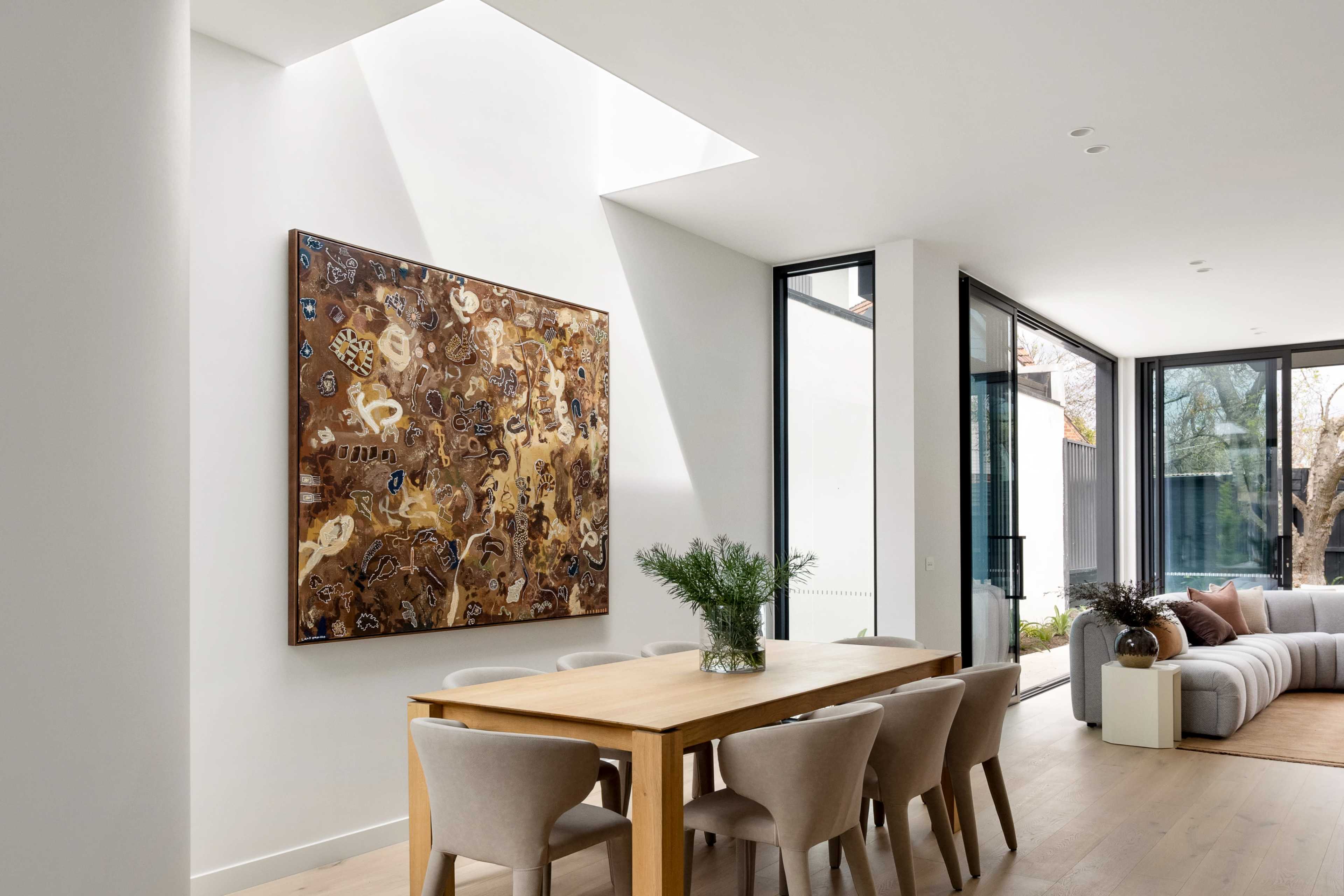 A modern dining area features a wooden table surrounded by upholstered chairs, with a large artwork displayed on the wall and natural light streaming in through a skylight.