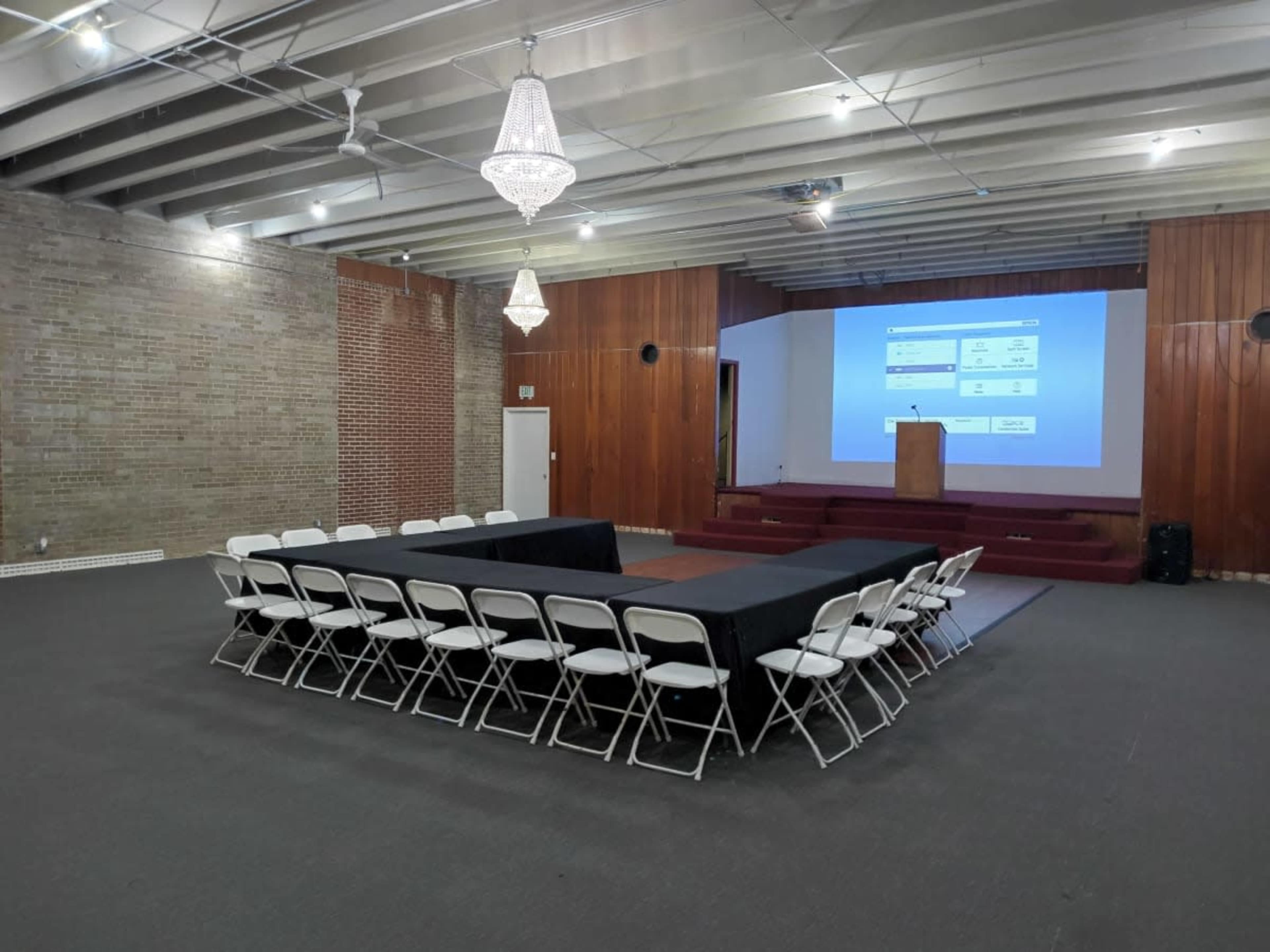 A rectangular arrangement of tables with chairs surrounds a small stage featuring a podium and a large screen in a spacious, well-lit room.