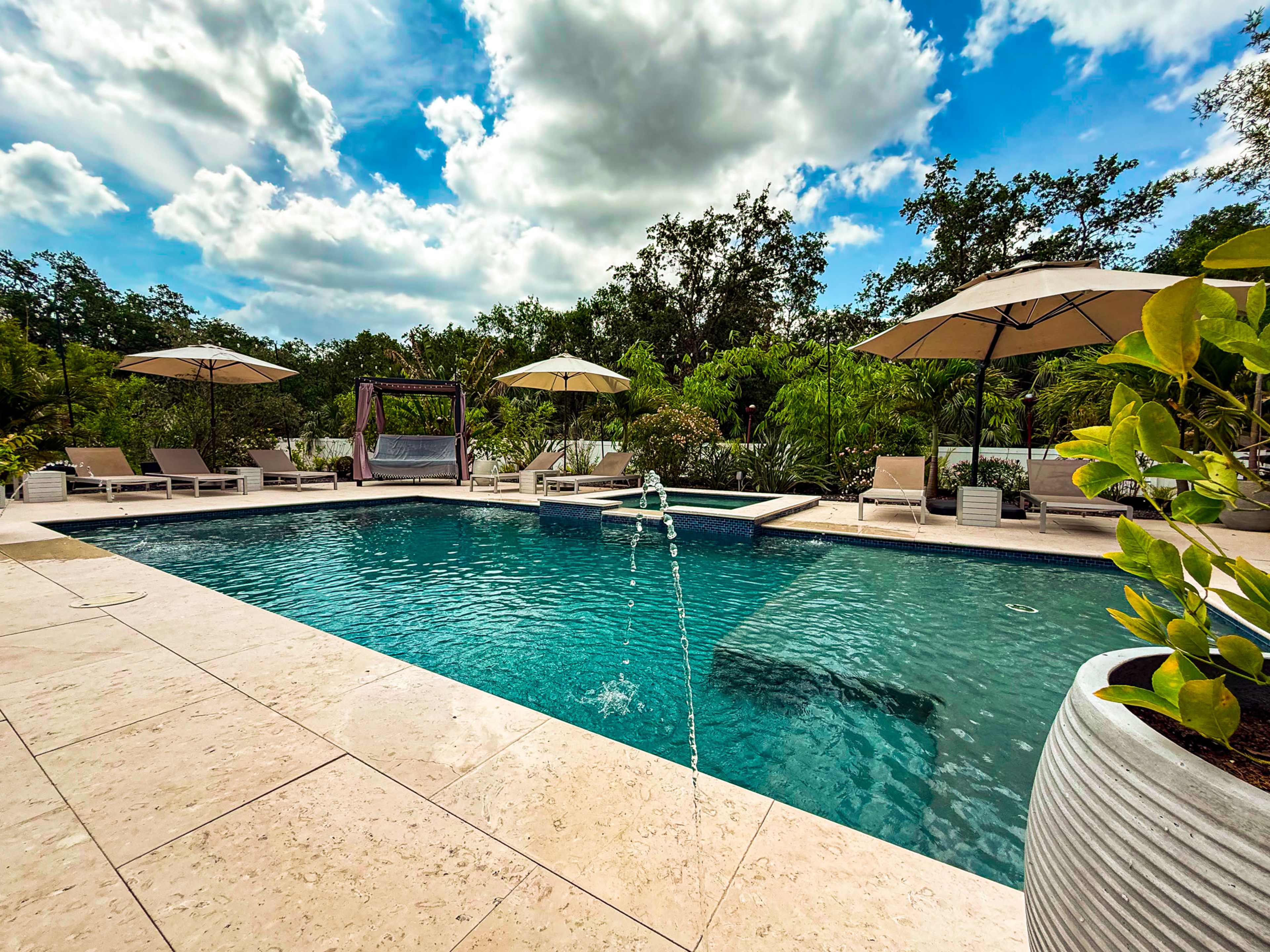 A backyard swimming pool with lounge chairs, umbrellas, and greenery surrounds it under a partly cloudy sky.