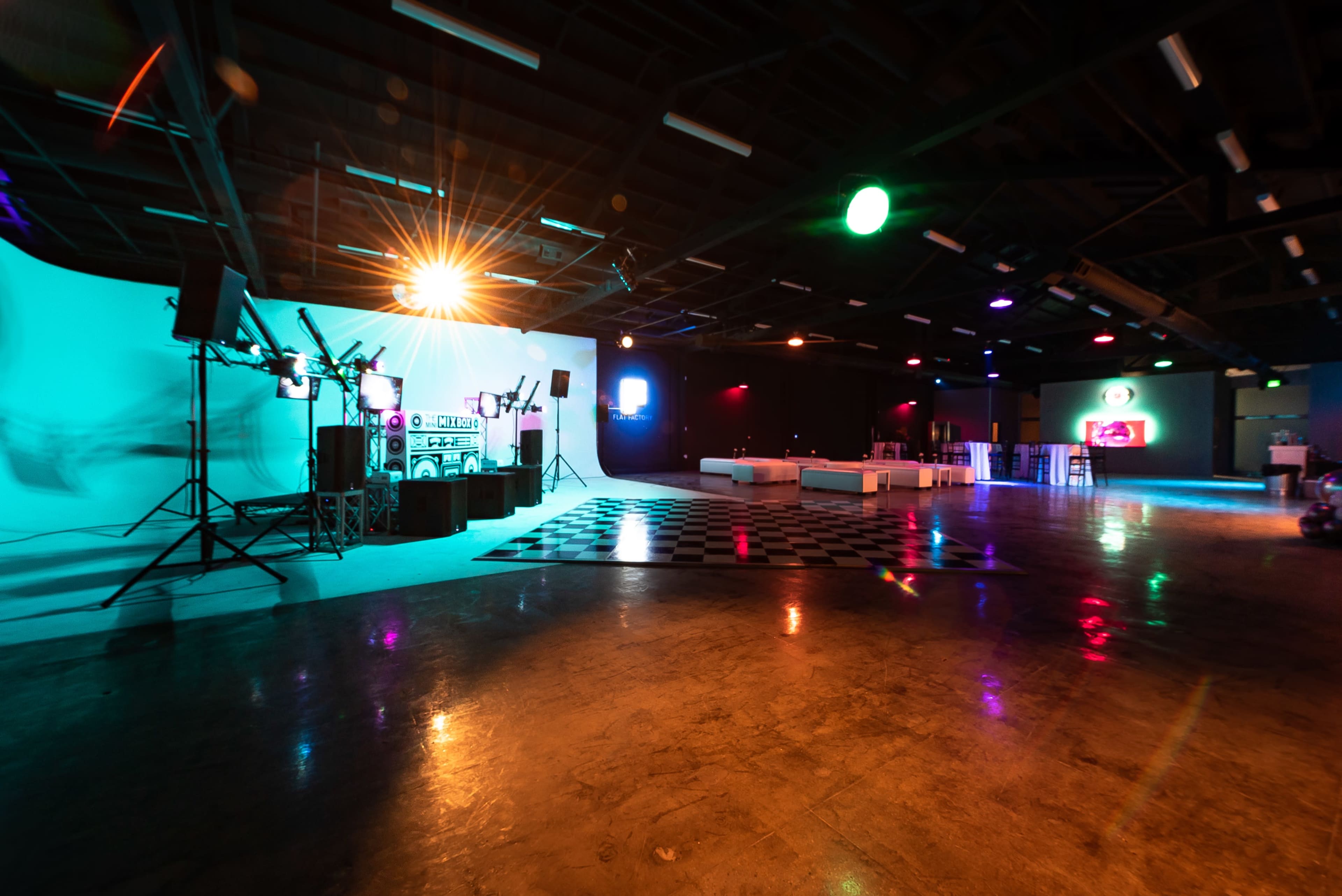 The image shows a spacious event venue with a checkered dance floor, colorful stage lighting, and various sound equipment set up for a gathering.
