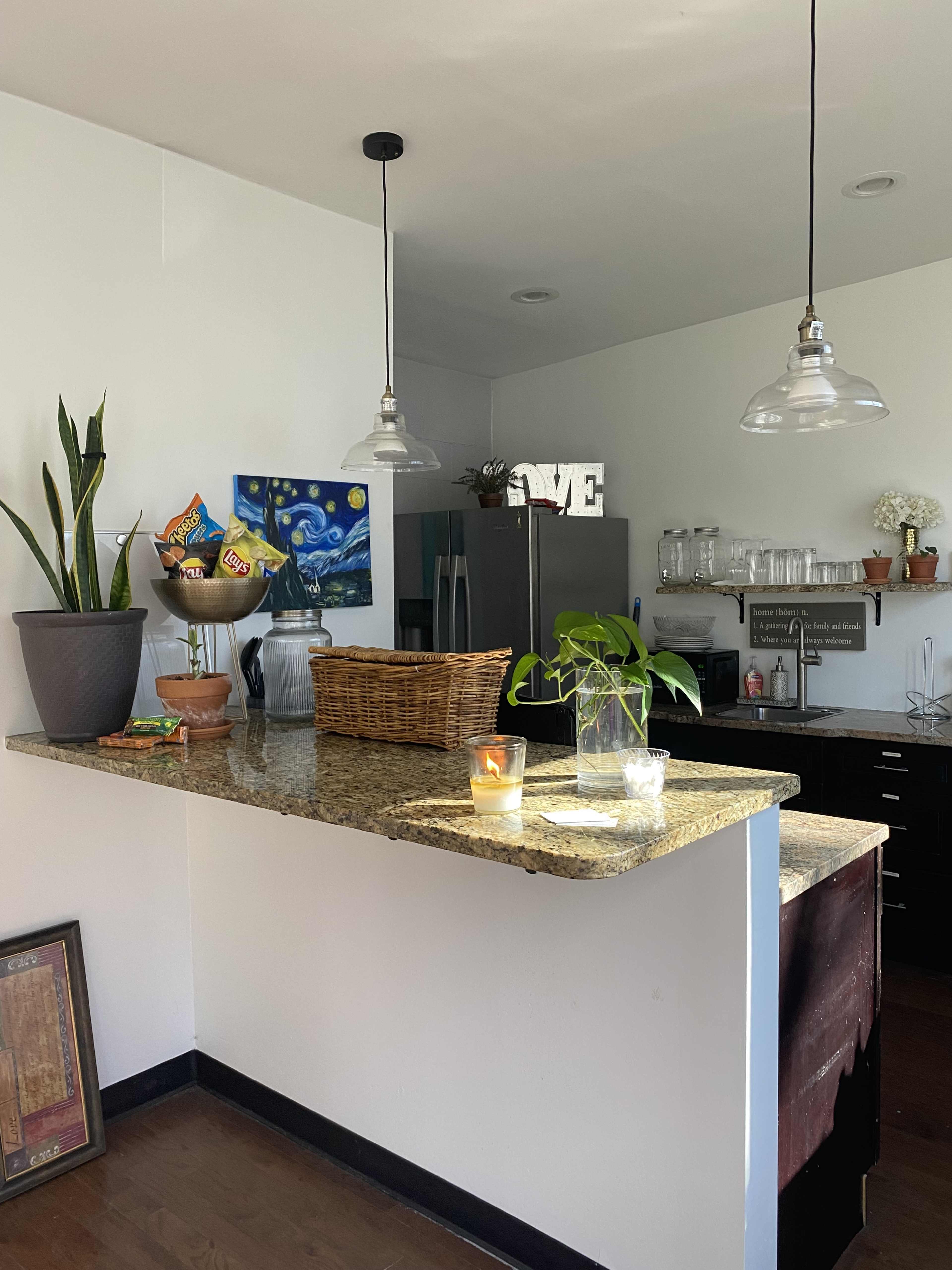 A granite kitchen island is adorned with a basket of snacks, a potted plant, and a candle, while overhead lights illuminate the space.