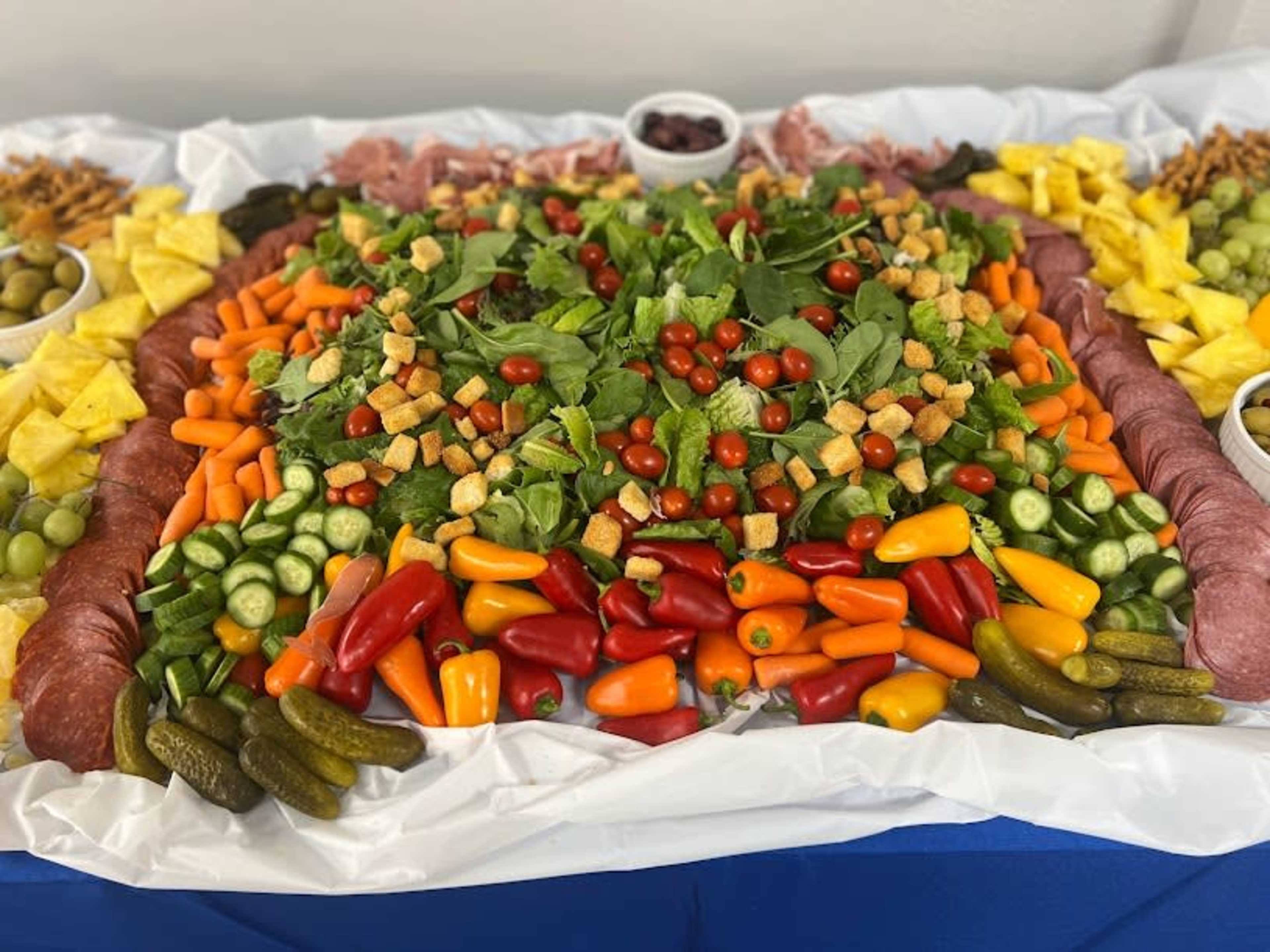 A large platter is arranged with an assortment of salad greens, cherry tomatoes, assorted peppers, pickles, olives, and various fruits, surrounded by bowls of grapes and crackers.