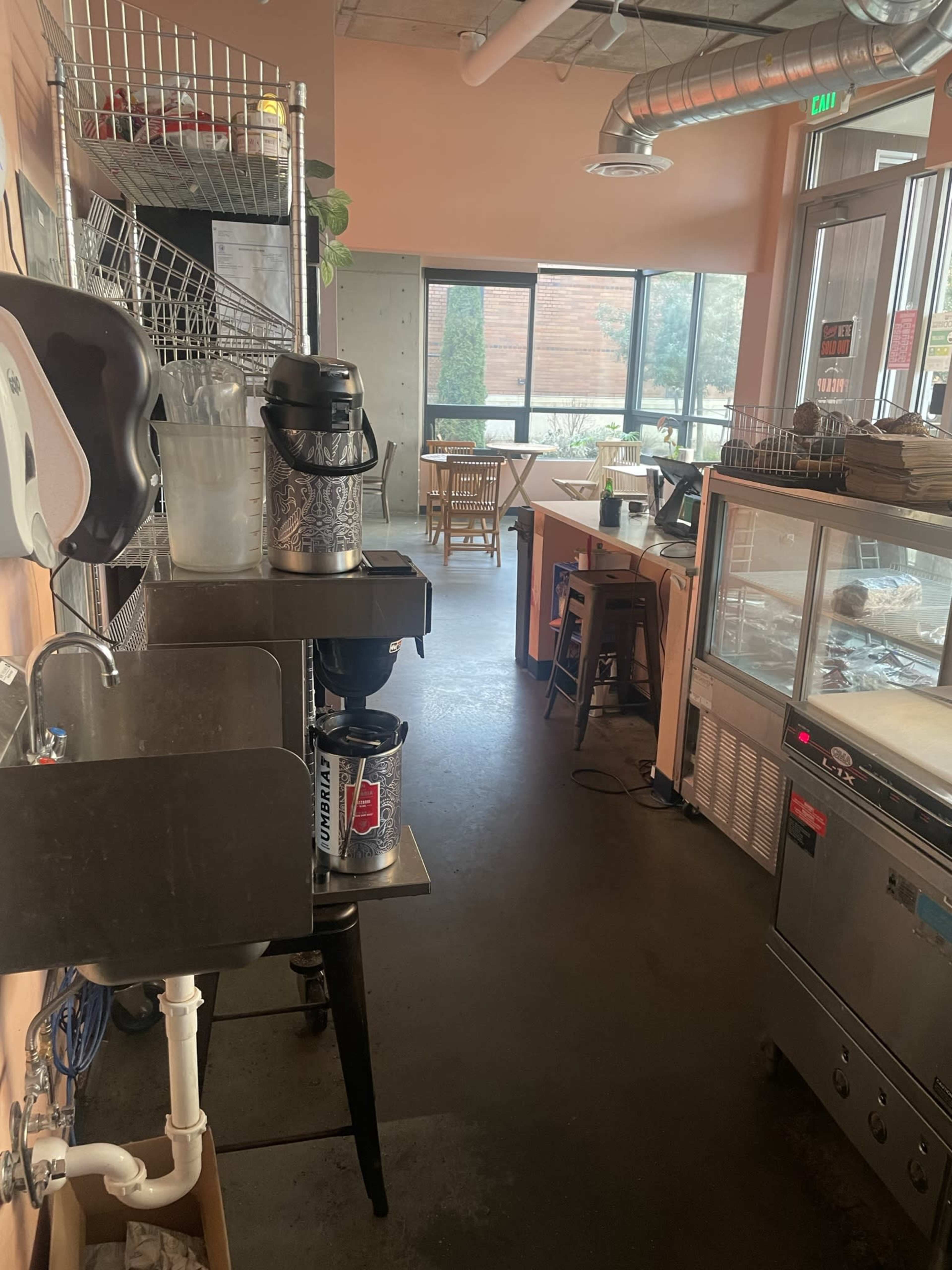 The image shows the interior of a café with a clean workspace, including a sink and beverage equipment, leading to a dining area with tables and large windows.