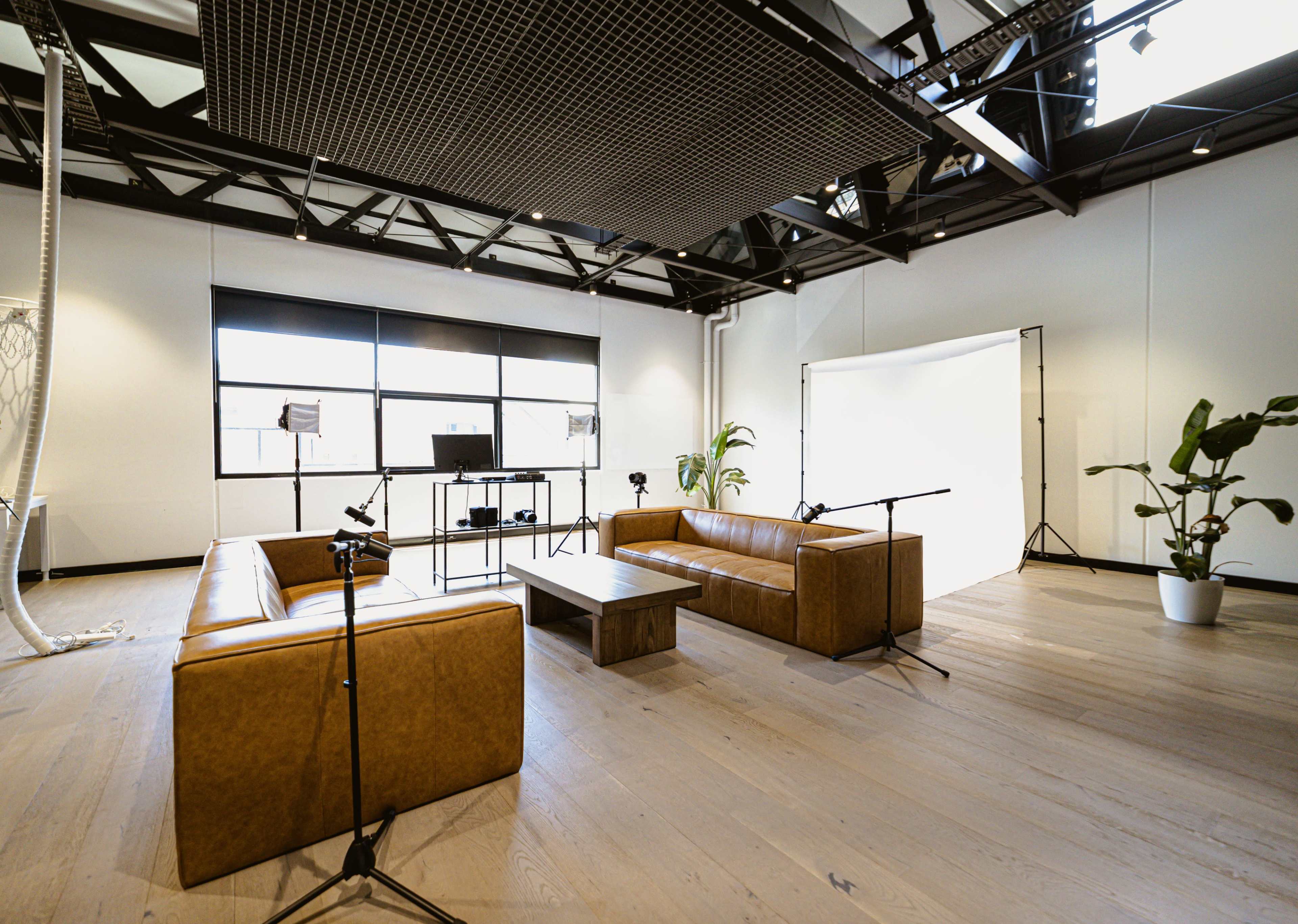 A modern studio space with two brown leather sofas, a coffee table, and lighting equipment arranged near a plain backdrop.