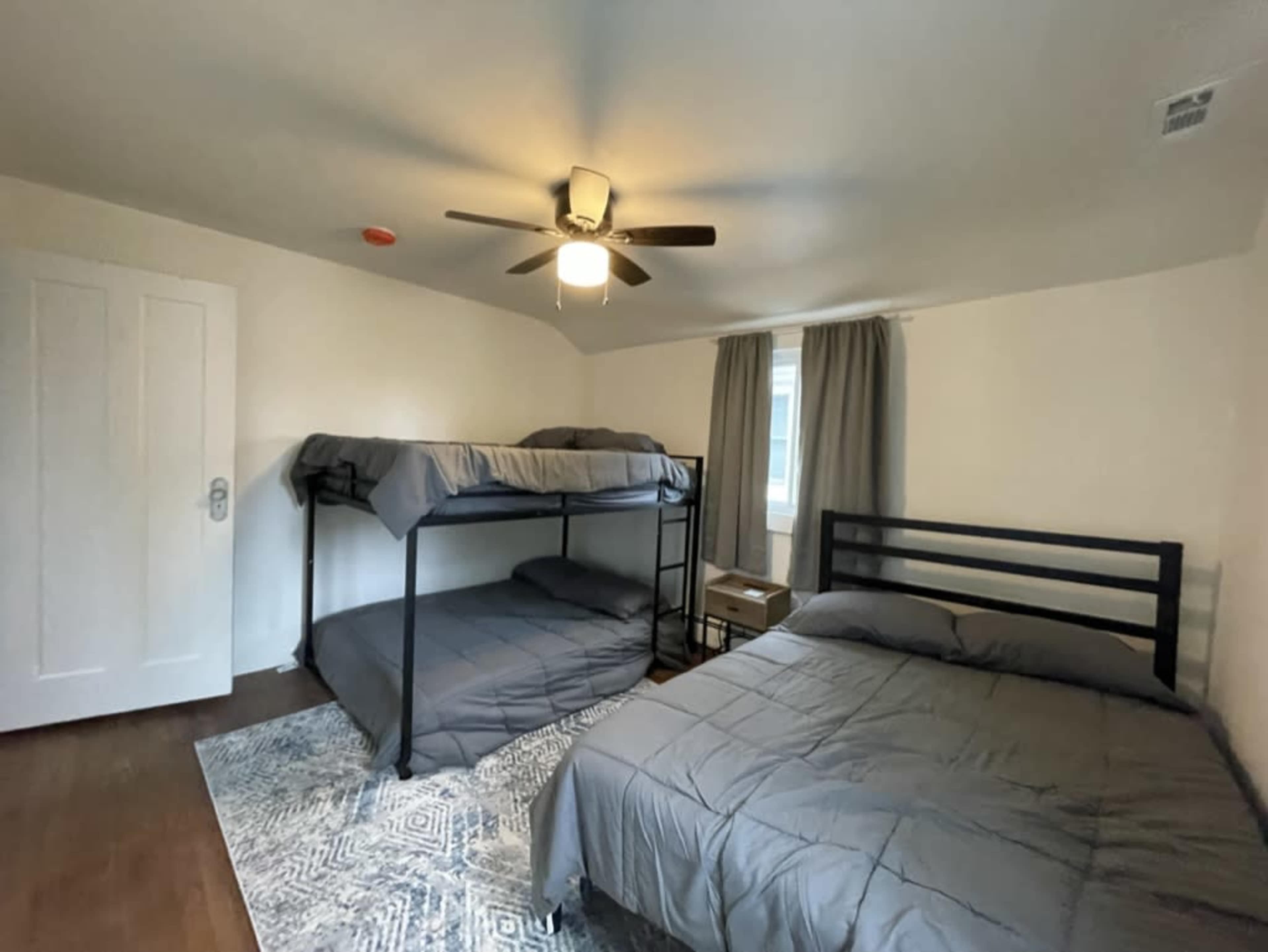 The image shows a bedroom featuring a bunk bed and a separate bed, with a ceiling fan and two windows covered by curtains.