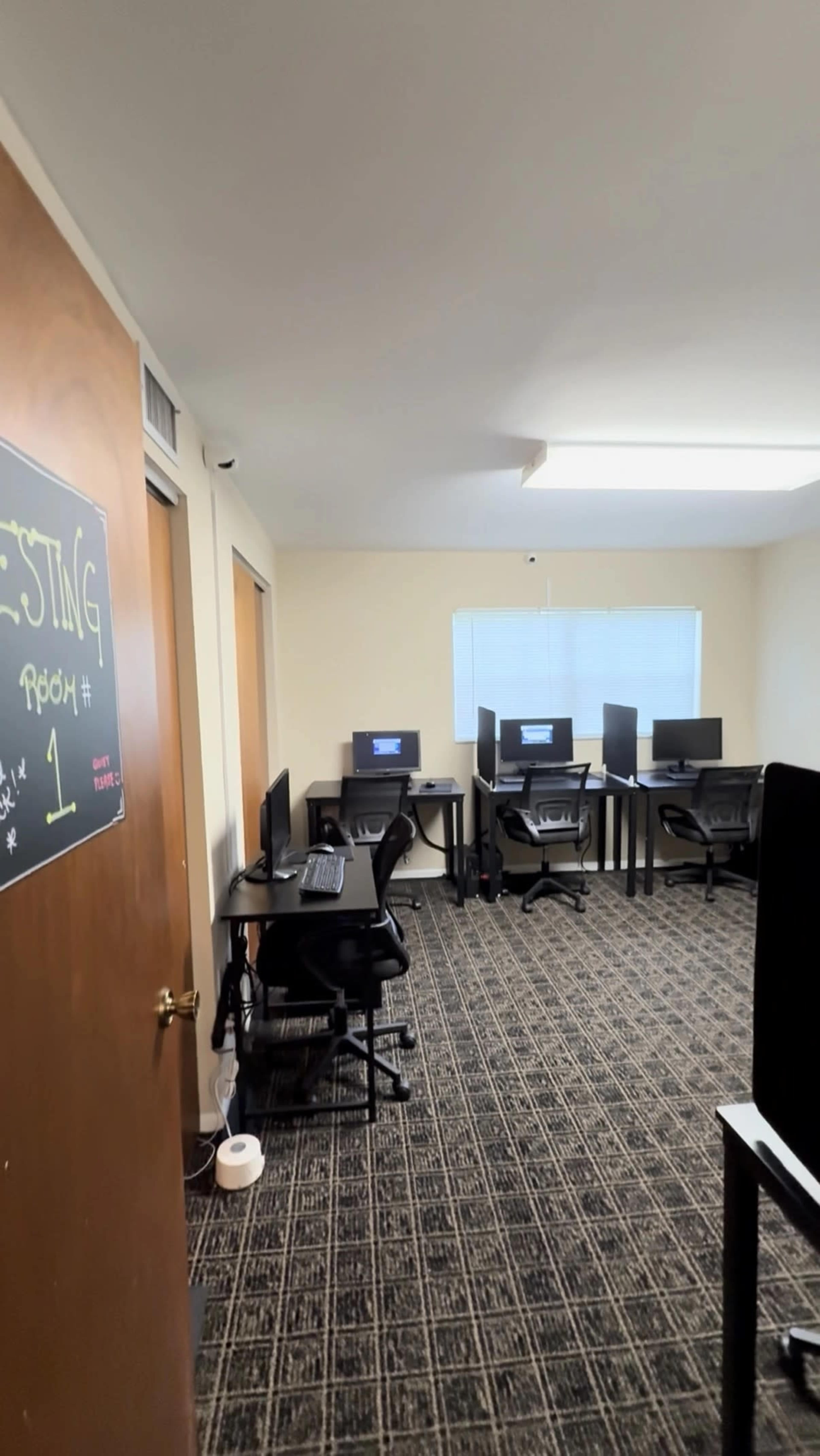 The image shows a small office space with several computer workstations arranged along the walls and a doorway labeled "Testing Room #1."