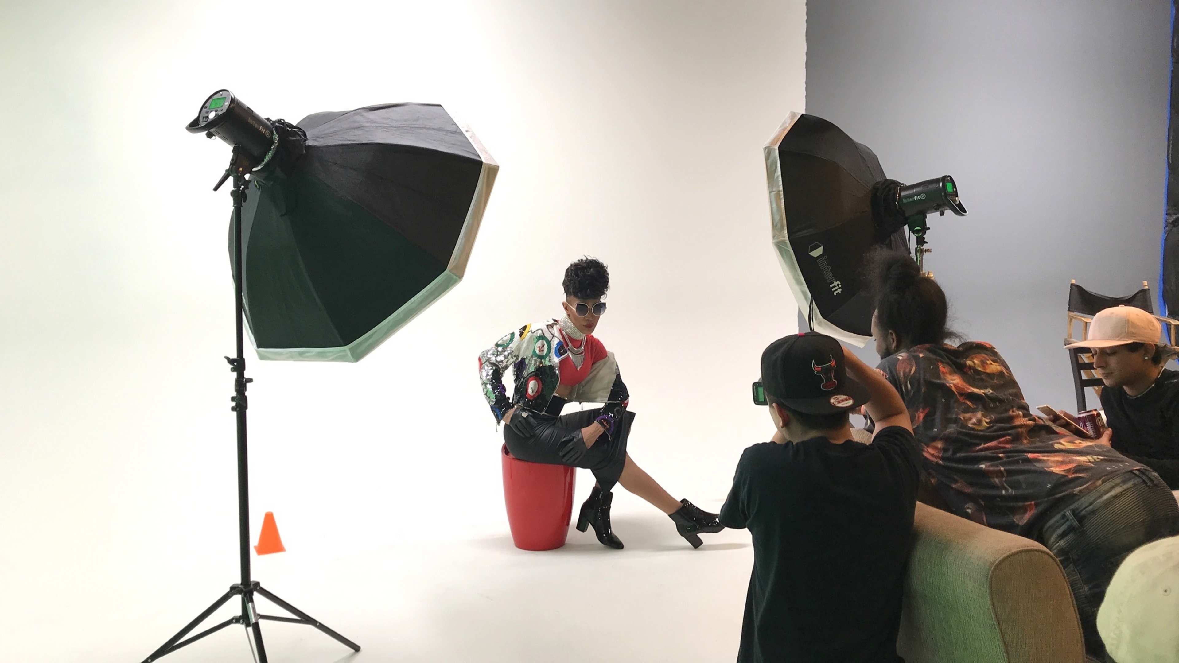 A model poses on a red stool in front of a white backdrop while photographers adjust lighting equipment.