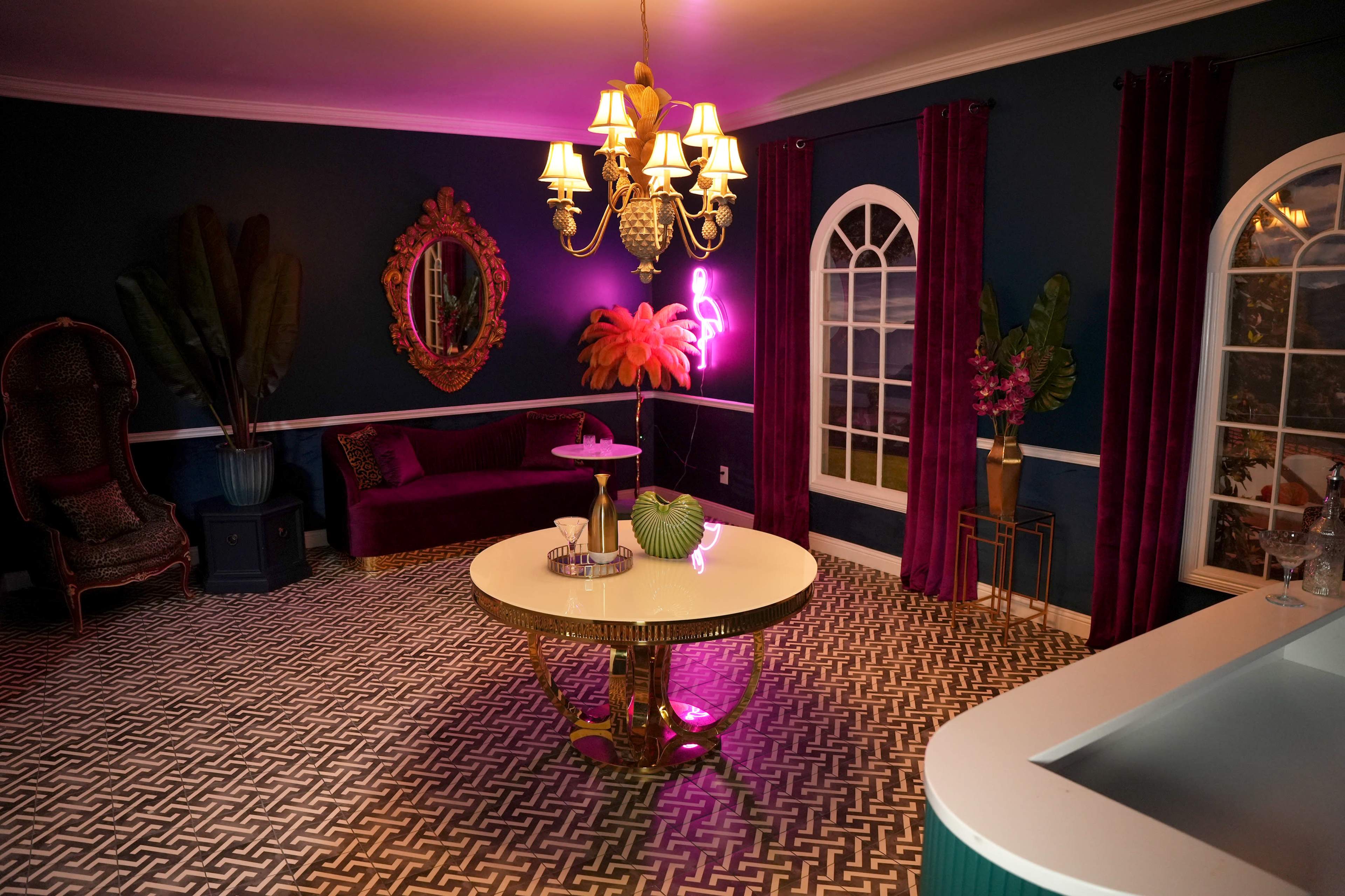 The image shows a stylish indoor living space featuring a dark teal wall, a round white table with gold accents, a plush purple couch, and decorative plants alongside a vibrant chandelier.