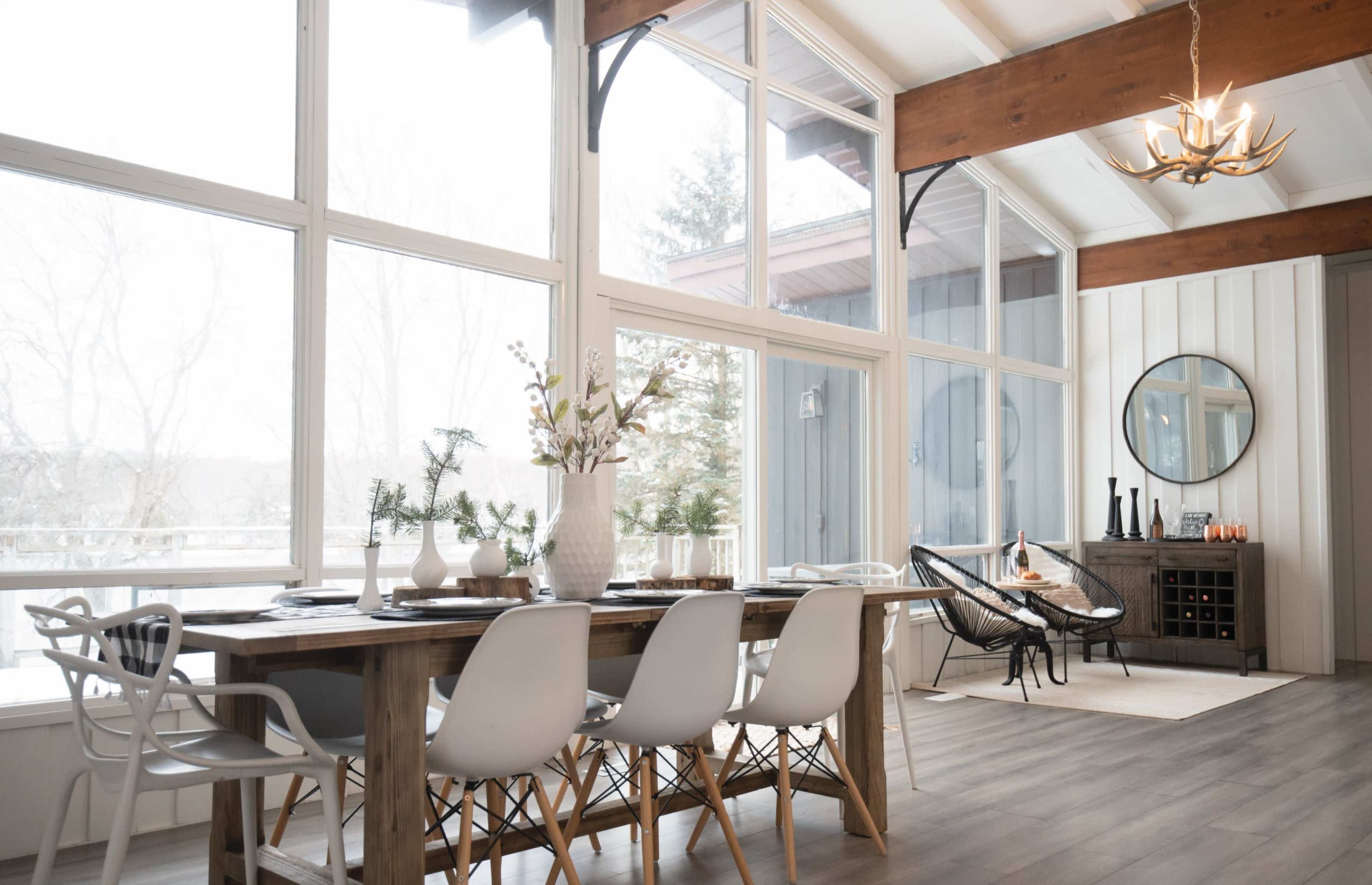 A bright dining area featuring a wooden table with white and light grey chairs, large windows, and a decorative circular mirror on the wall.