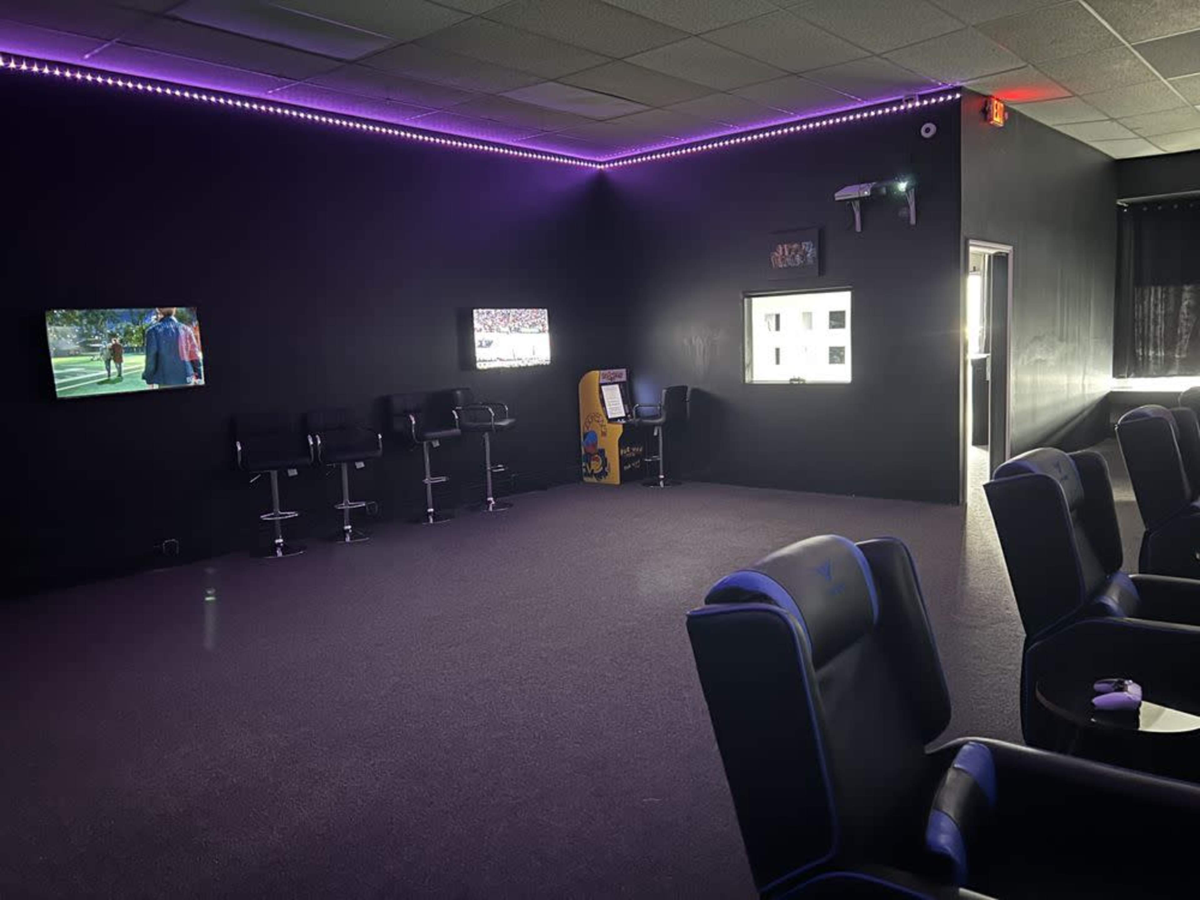 The image shows a dark room with two televisions on the walls, seating in the form of black chairs, and an arcade game console in one corner.