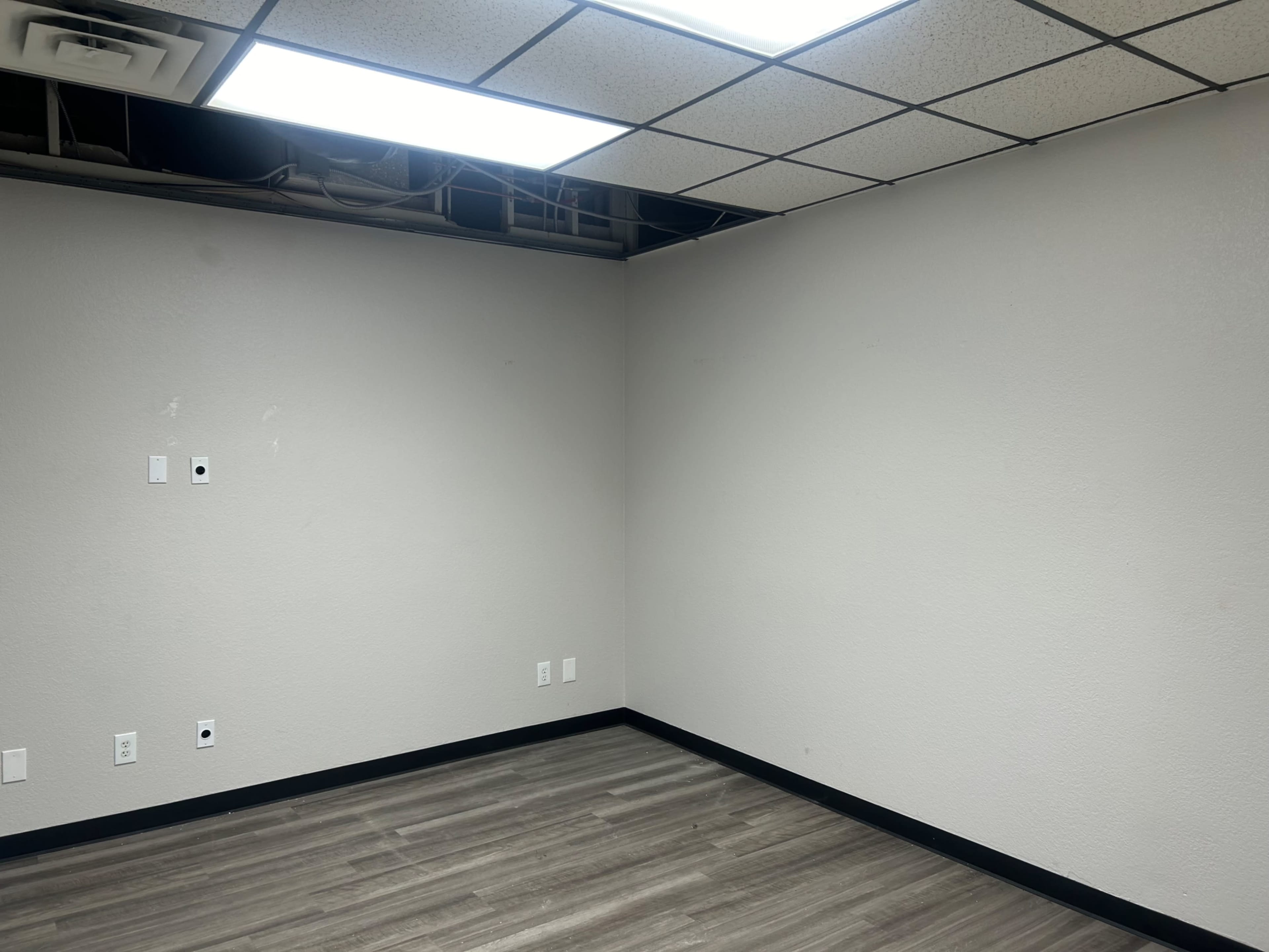 The image shows a sparsely furnished room with beige walls, a black trim along the floor, and a partially exposed ceiling.