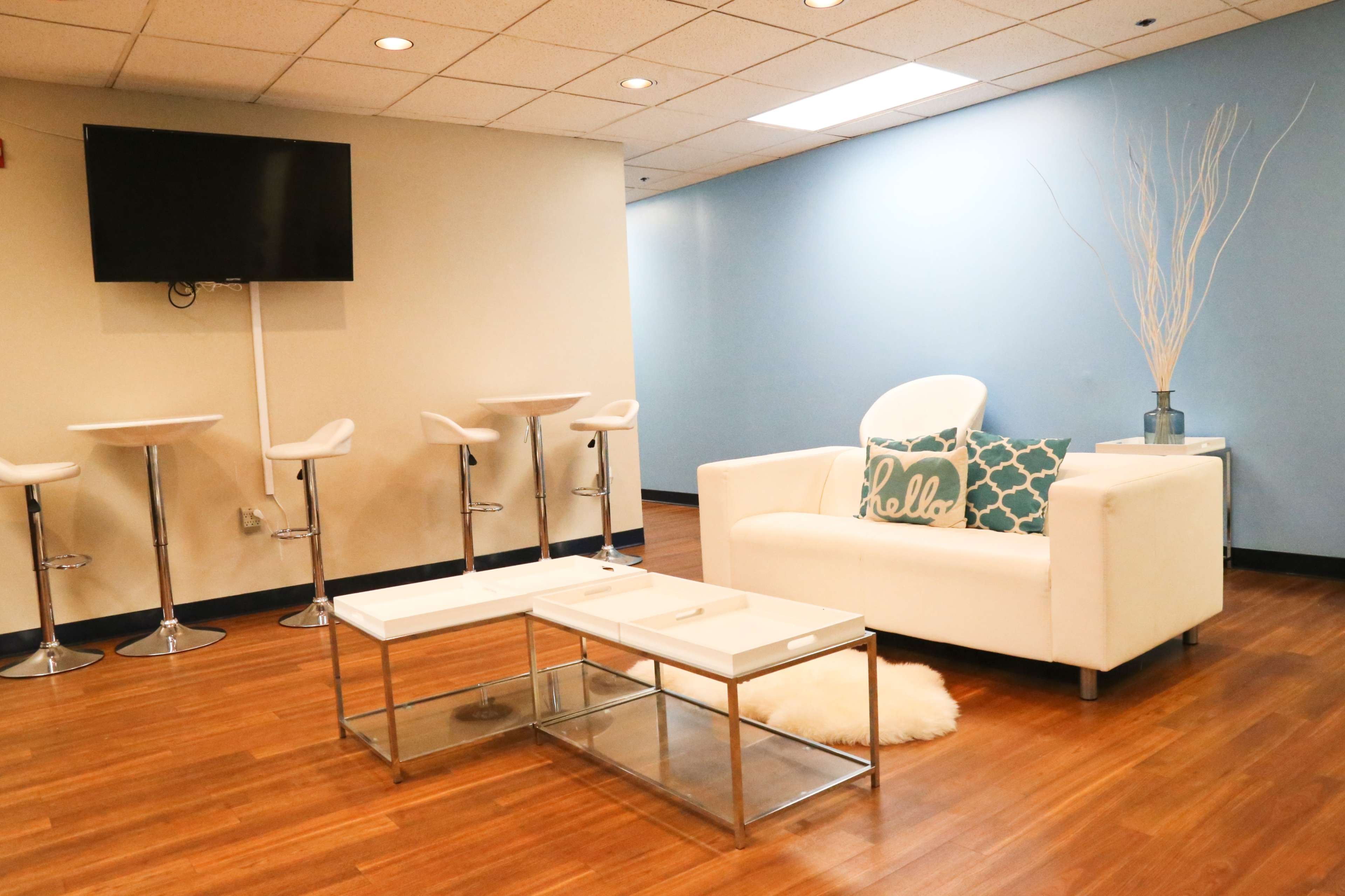 A modern lounge area with a white sofa, a coffee table, bar stools, and a television on the wall.