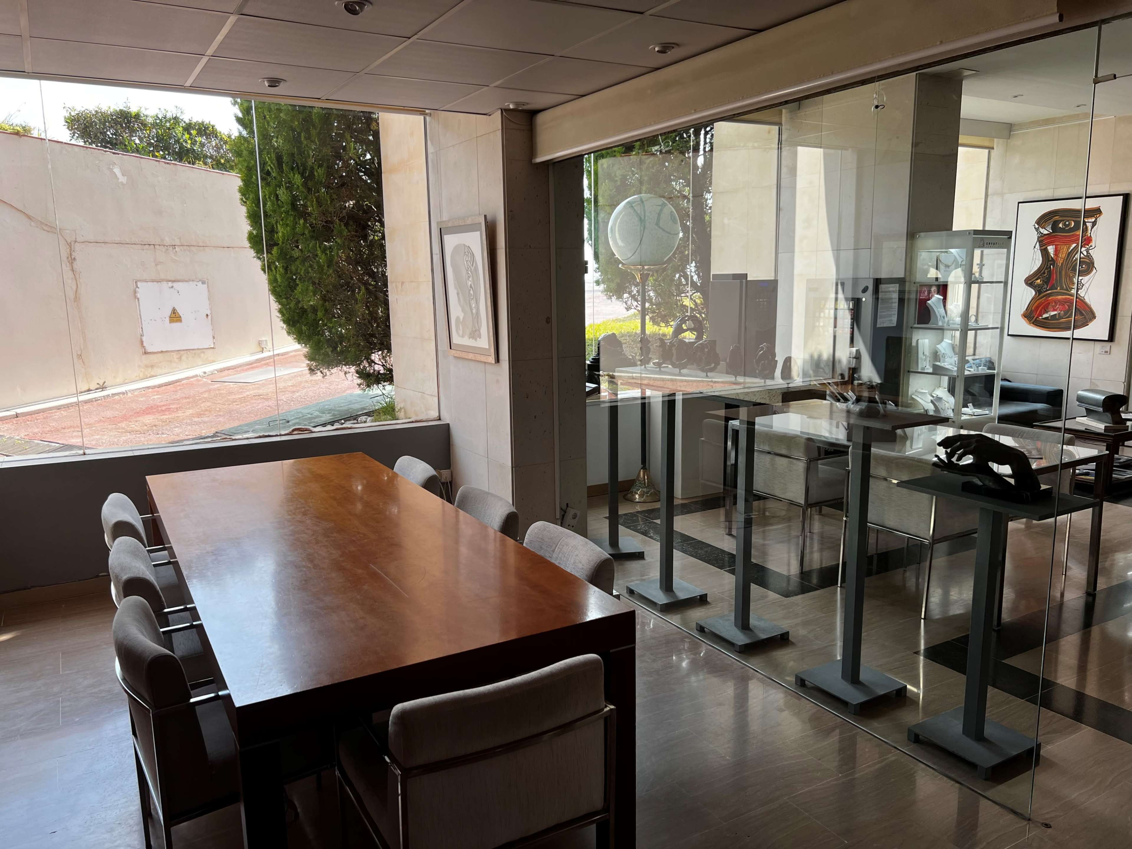 The image shows a modern conference room with a long wooden table and several chairs, adjacent to a glass wall displaying artwork and a view of an outdoor area.