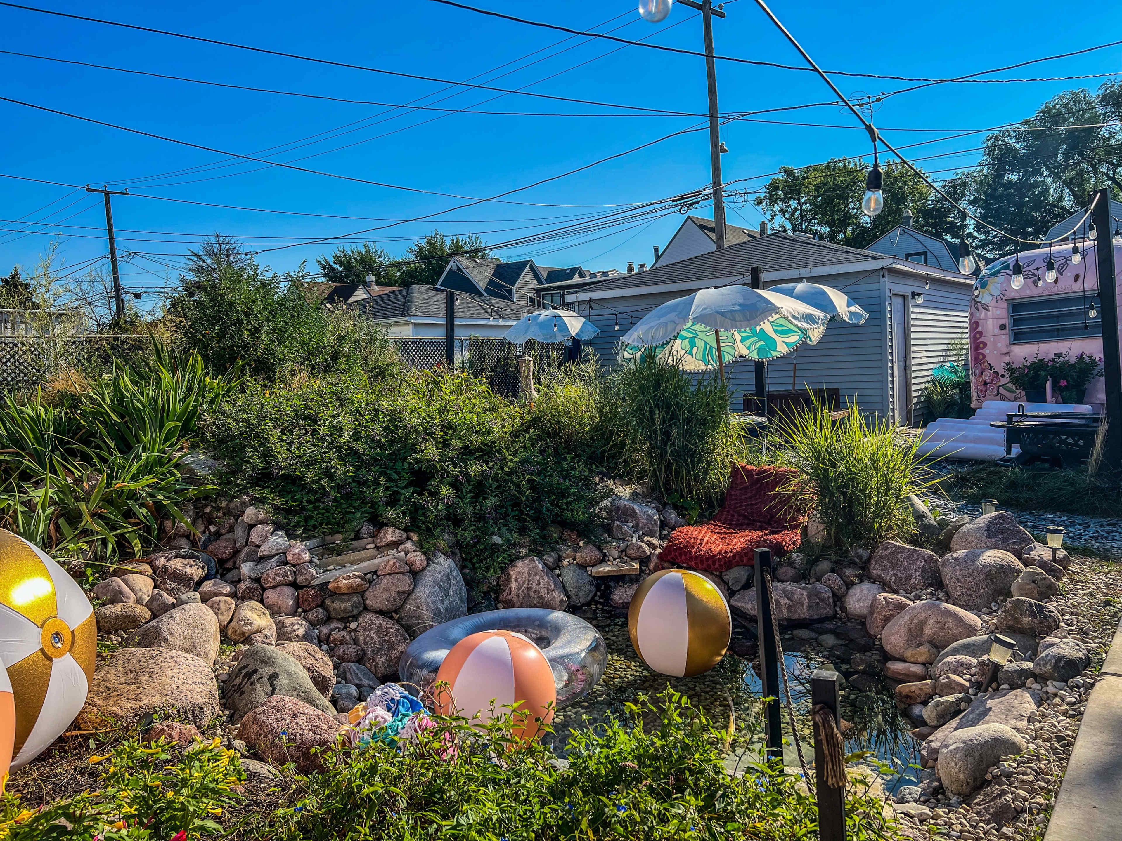 A backyard scene features a rock garden with beach balls, umbrellas, and string lights, set against a clear blue sky.