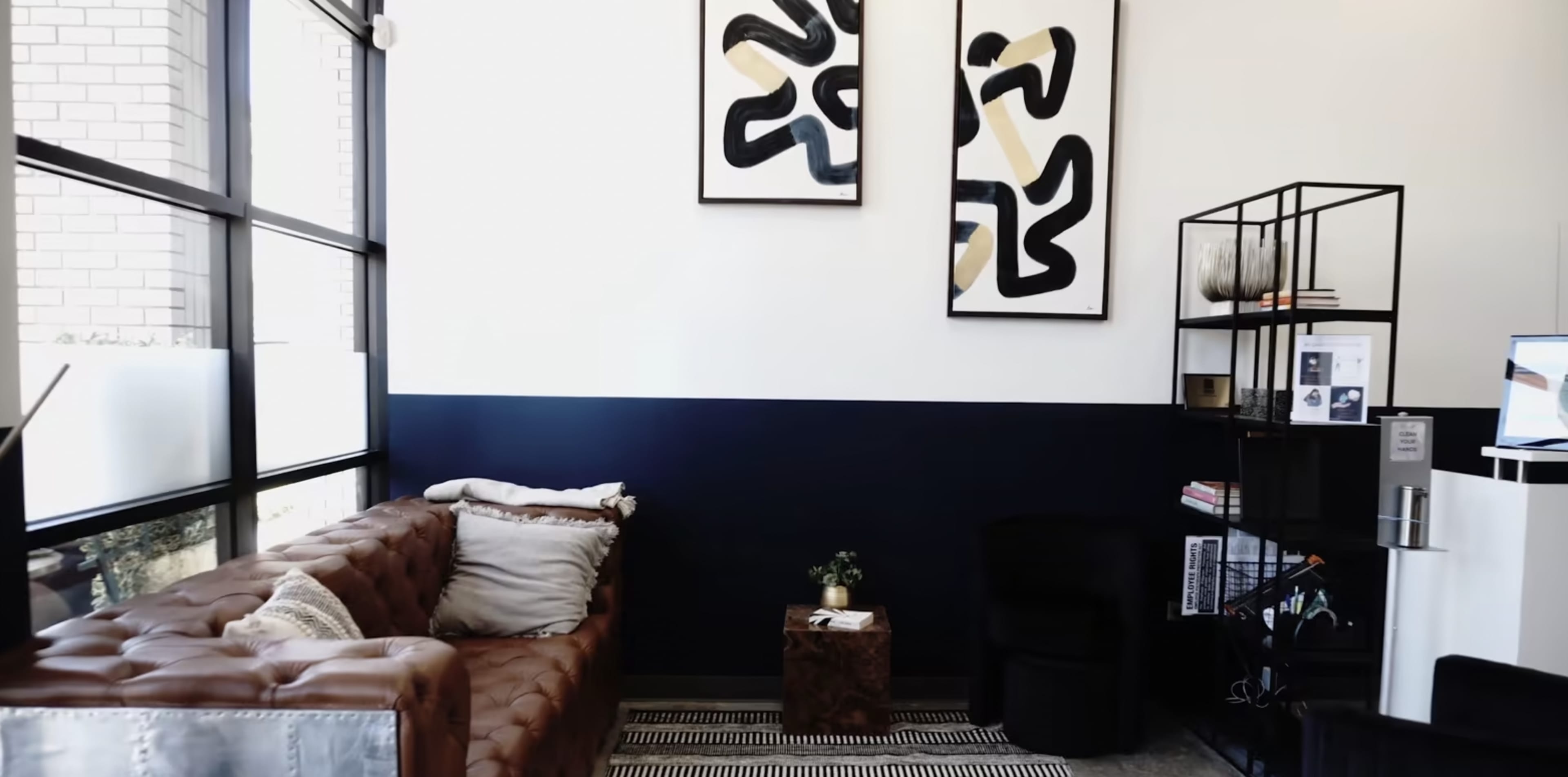 A modern interior with a brown leather sofa, two abstract art pieces on the wall, and a black bookshelf filled with items.