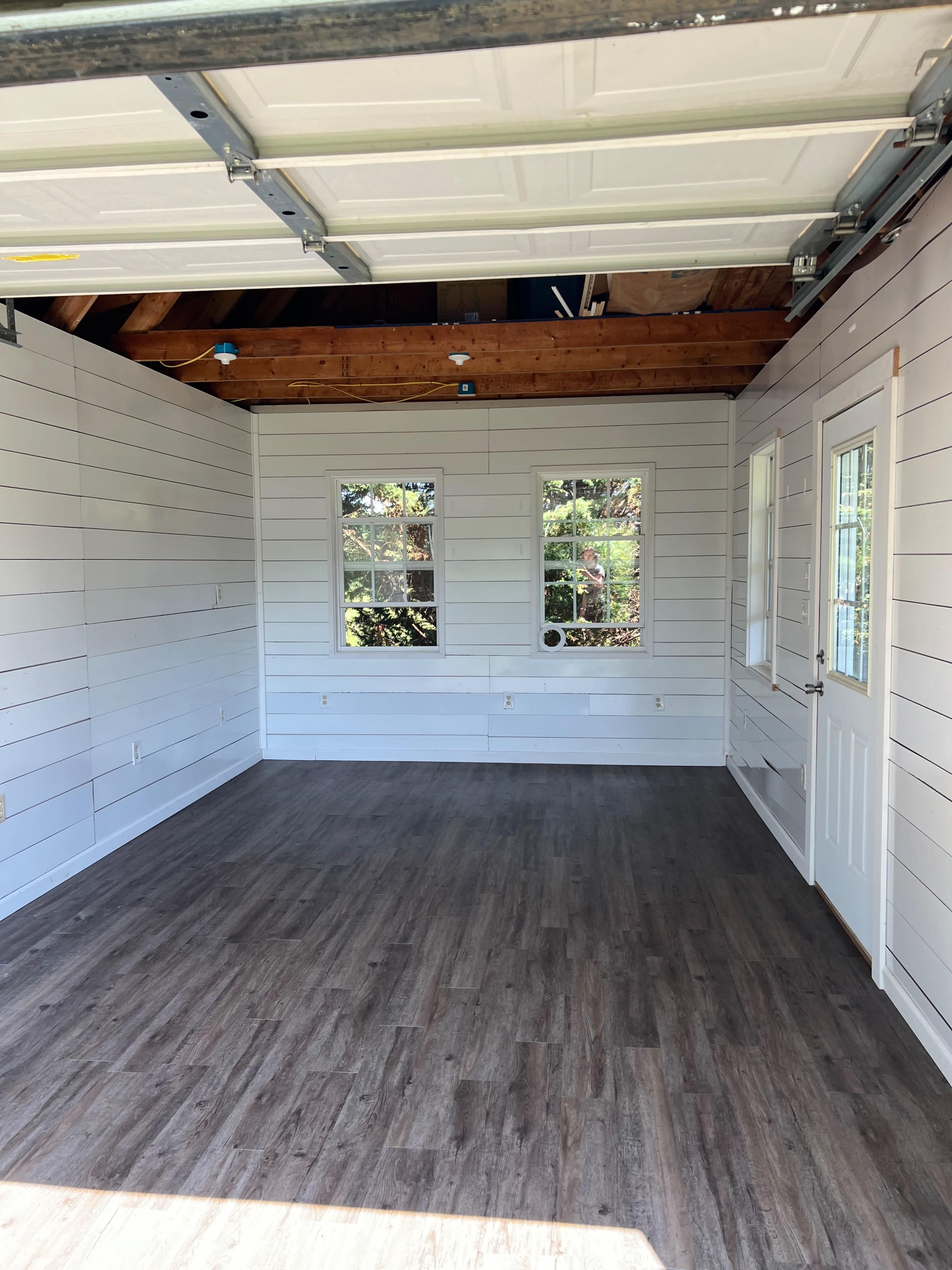 The image shows a spacious, empty room with shiplap walls, three windows, and a door, featuring a wooden floor.