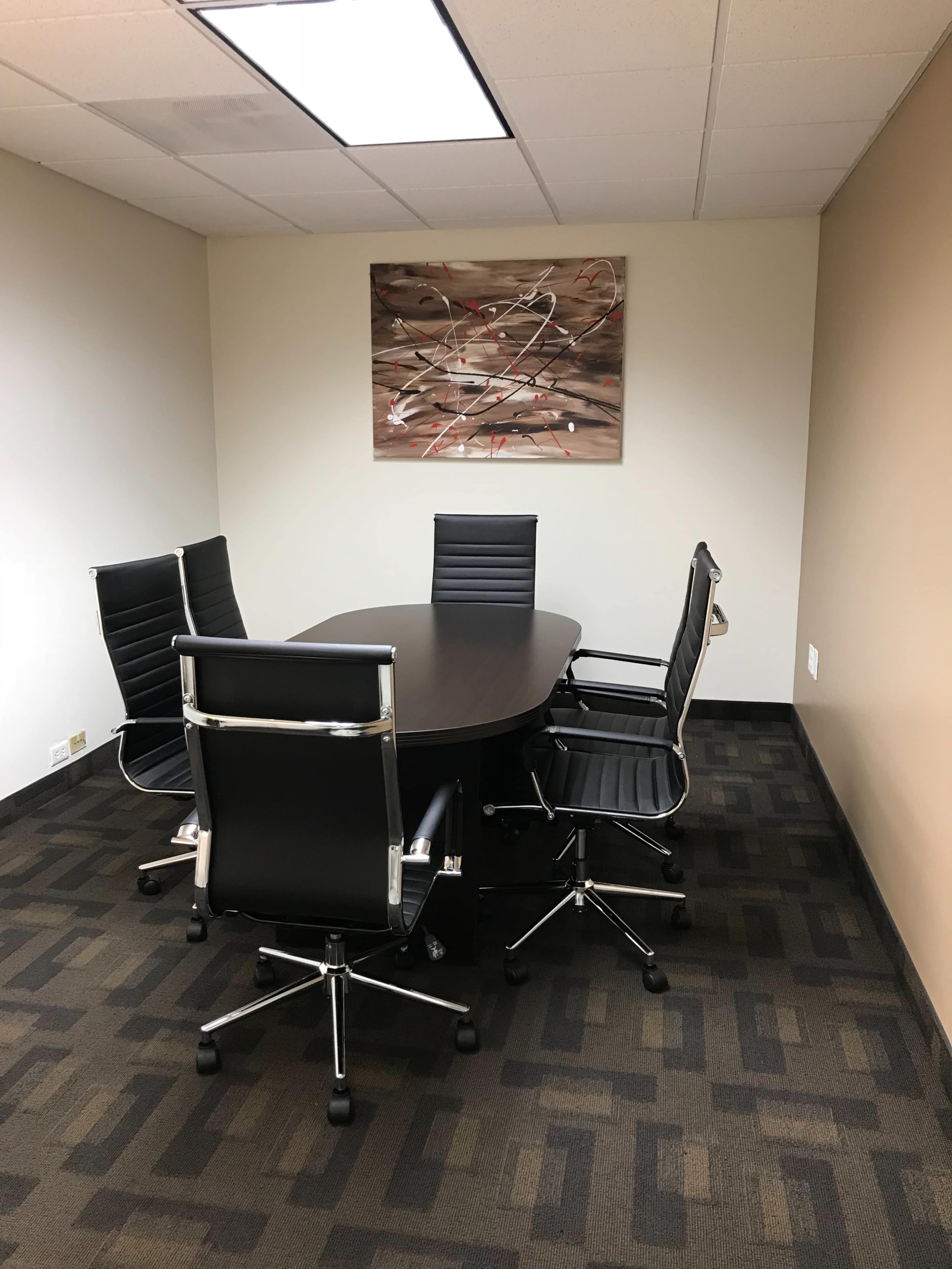 A conference room features a dark oval table surrounded by four black leather chairs, with a large abstract painting on the wall.