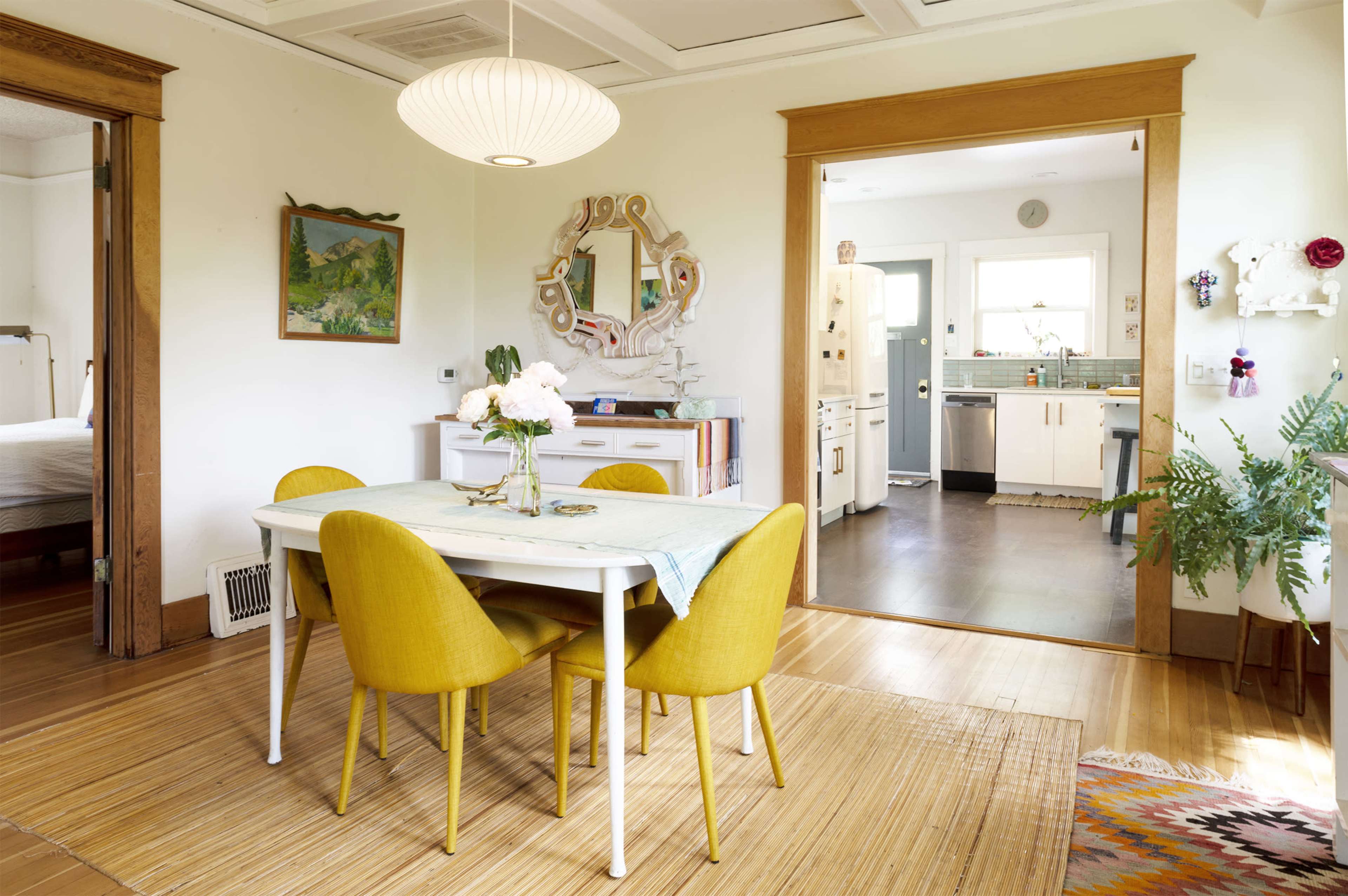 A dining area with a white table, four yellow chairs, and a vase of flowers is visible, leading to a kitchen through an open doorway.