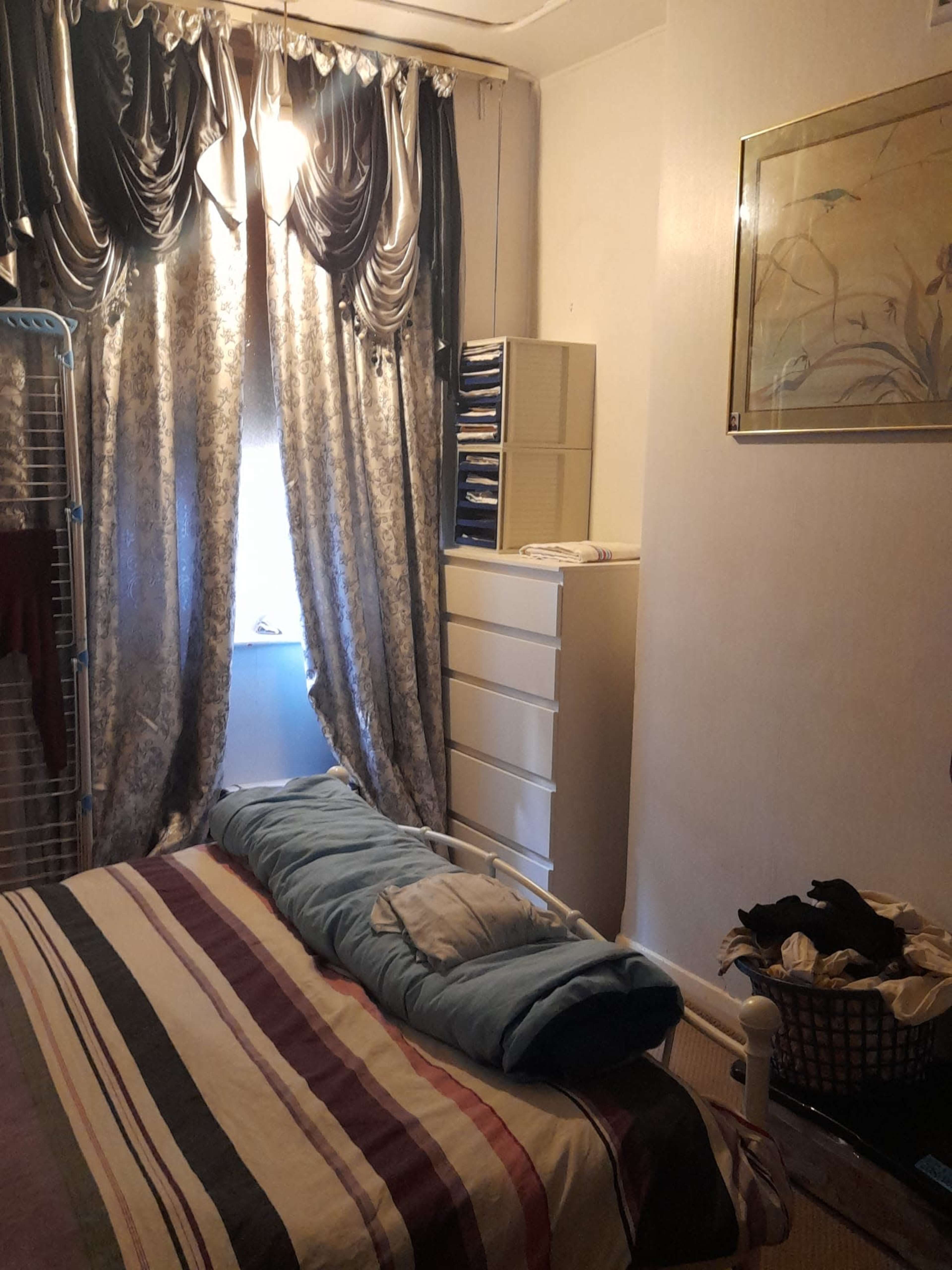 A small bedroom with a bed, a dresser, a window covered by heavy curtains, and clothes arranged on a drying rack.